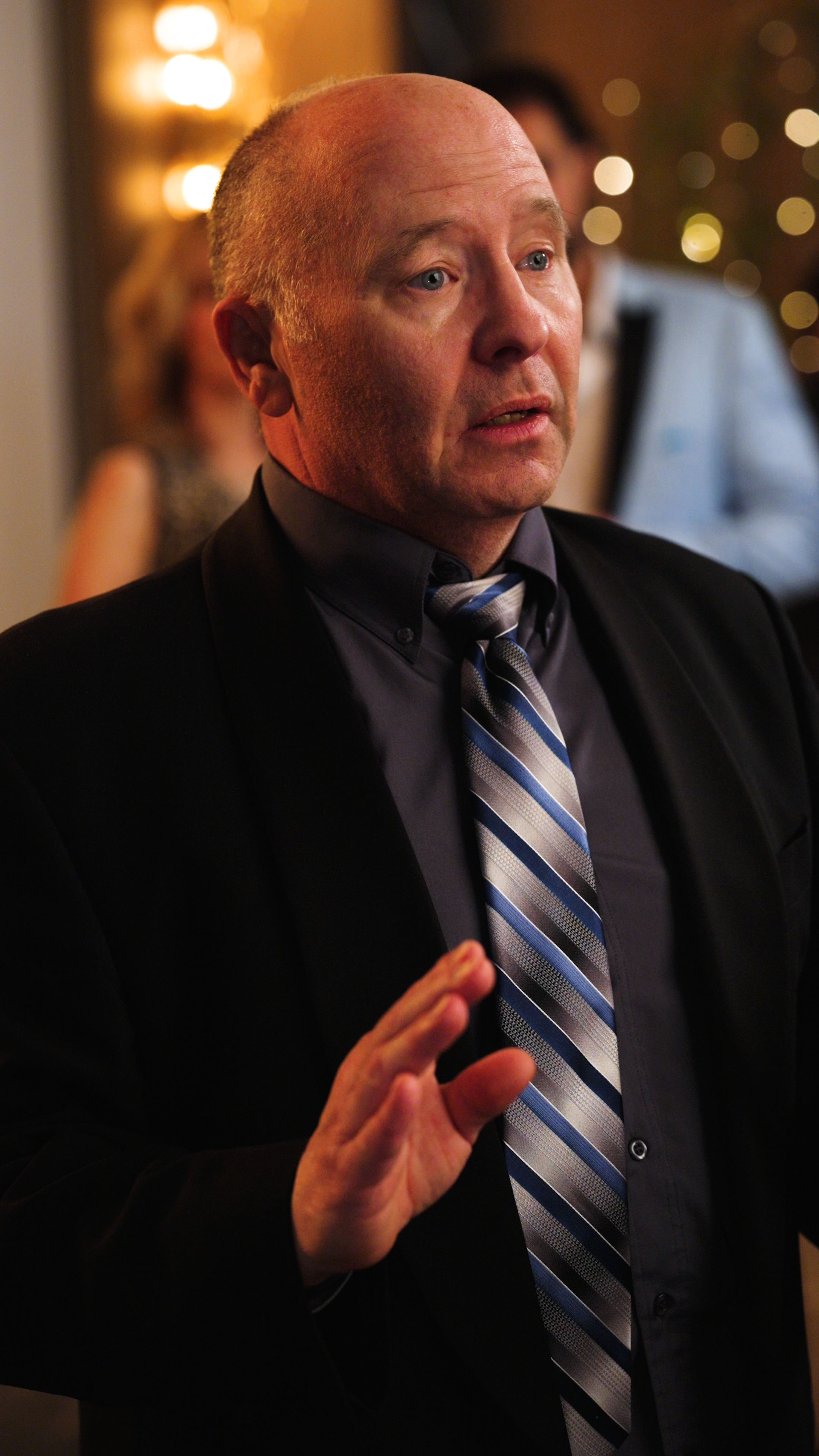 Middle-aged man in a black suit and striped tie raising his hand while speaking at a formal indoor event.