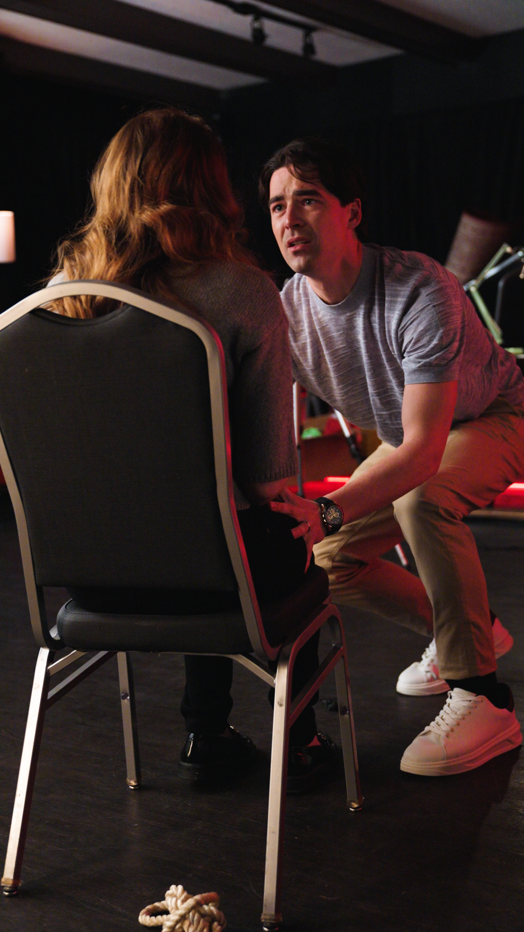 Man crouching in front of a woman seated on a chair with her hands behind her back under red lighting indoors.