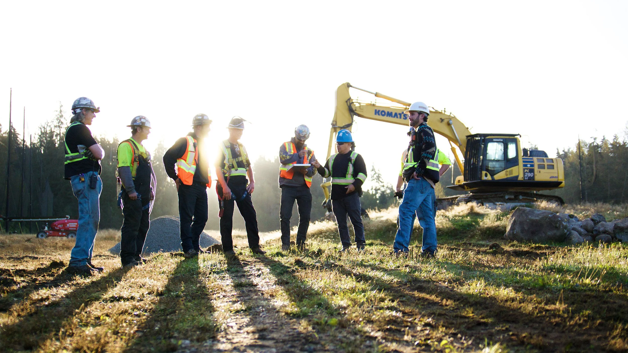 Construction crew meeting outdoors at a job site with an excavator in the background.