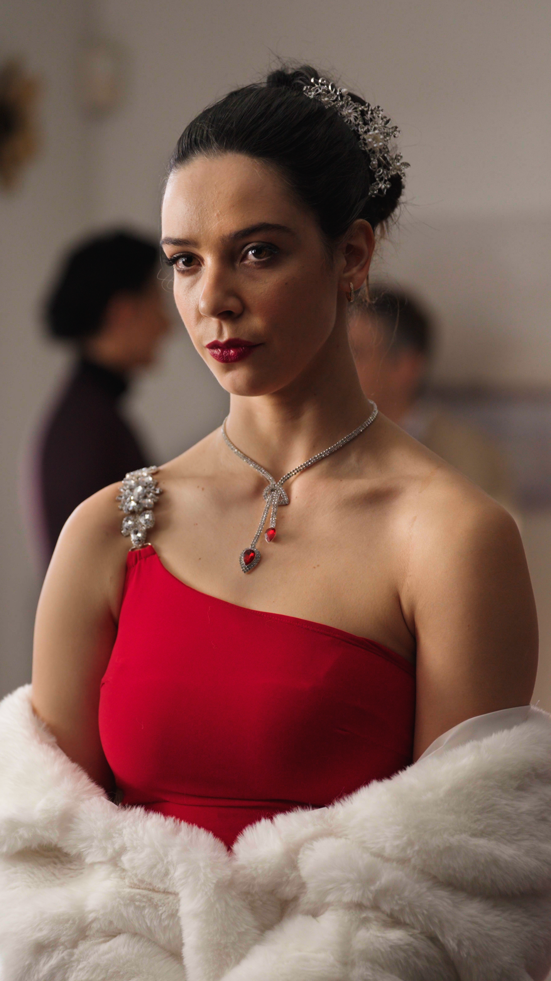 A woman stands poised in an elegant red one-shoulder gown, her dark hair styled in a sleek updo adorned with a delicate jeweled hairpiece. She wears a sparkling necklace with red gemstone accents that complement her dress, along with deep red lipstic