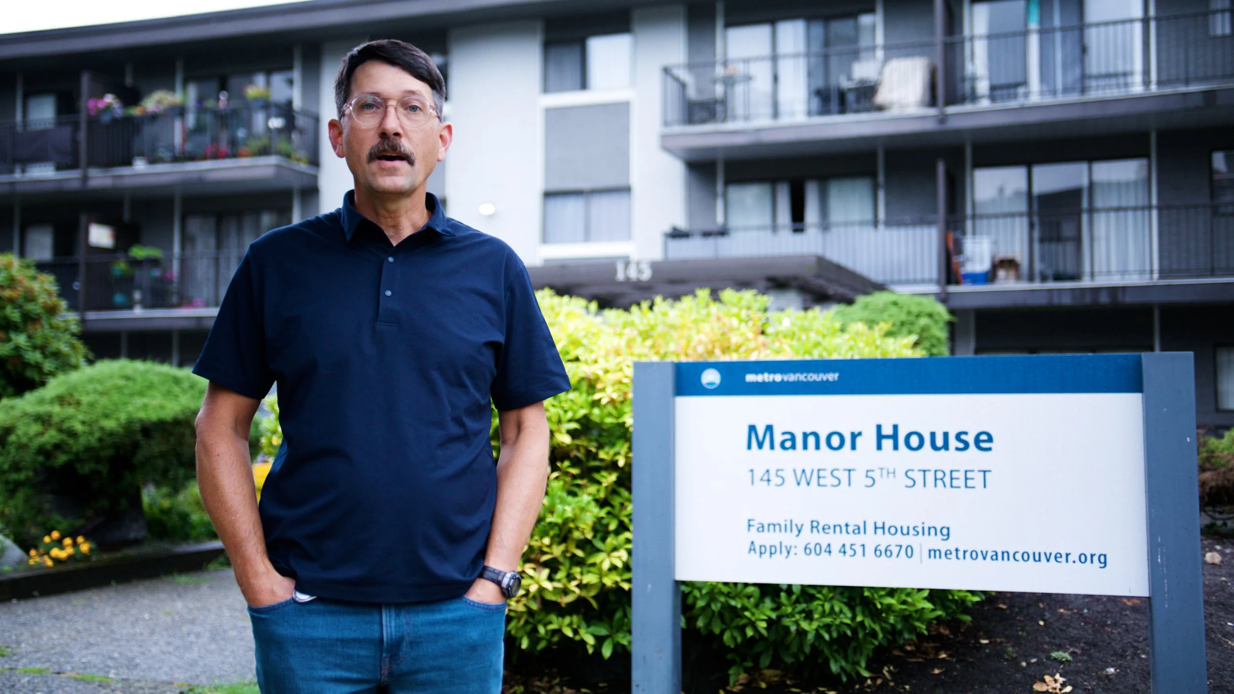 Man standing outside Manor House rental housing building in front of a Metro Vancouver sign.