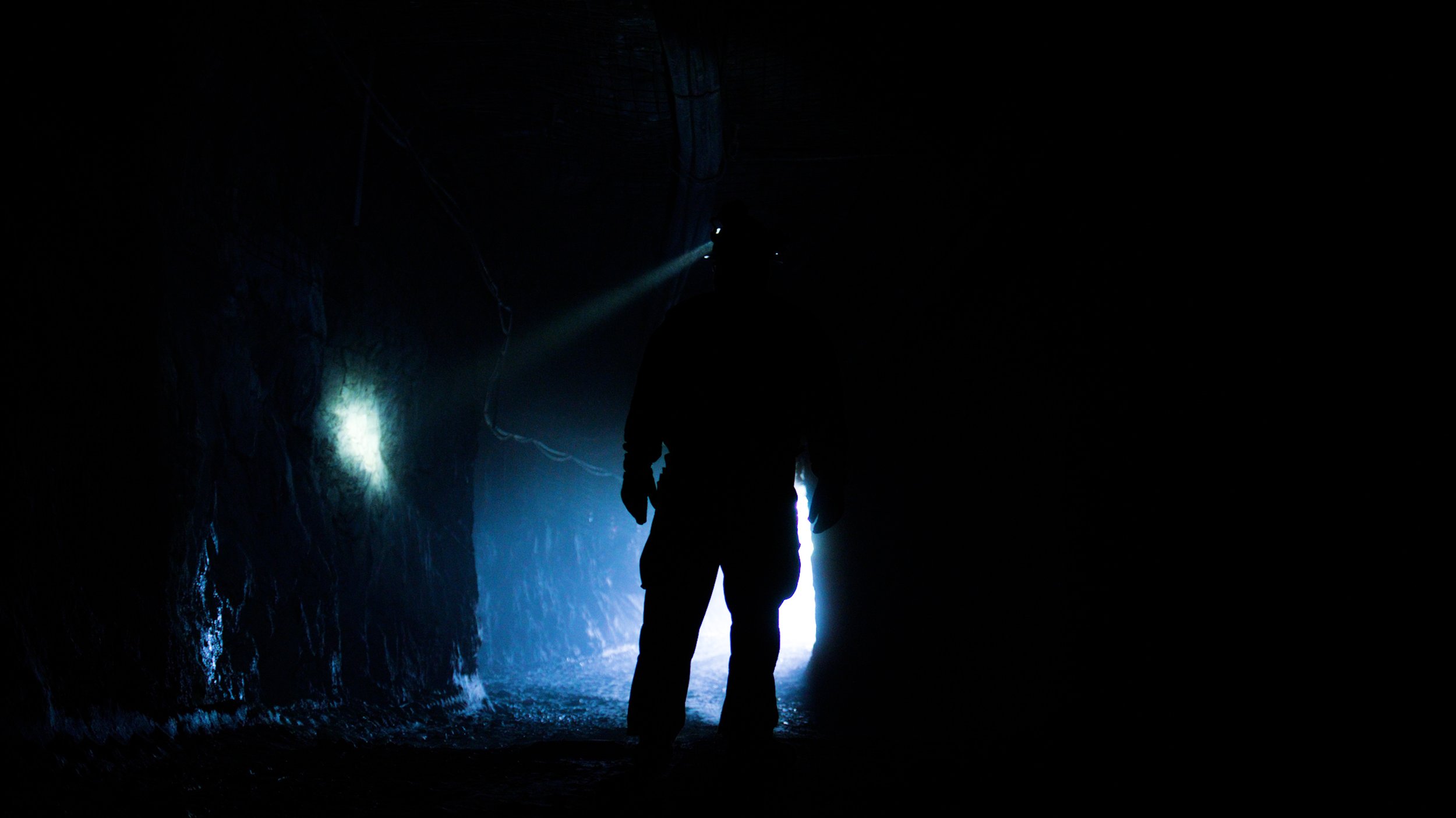 Silhouetted miner walking through an underground tunnel with headlamp lighting in a gold mine.