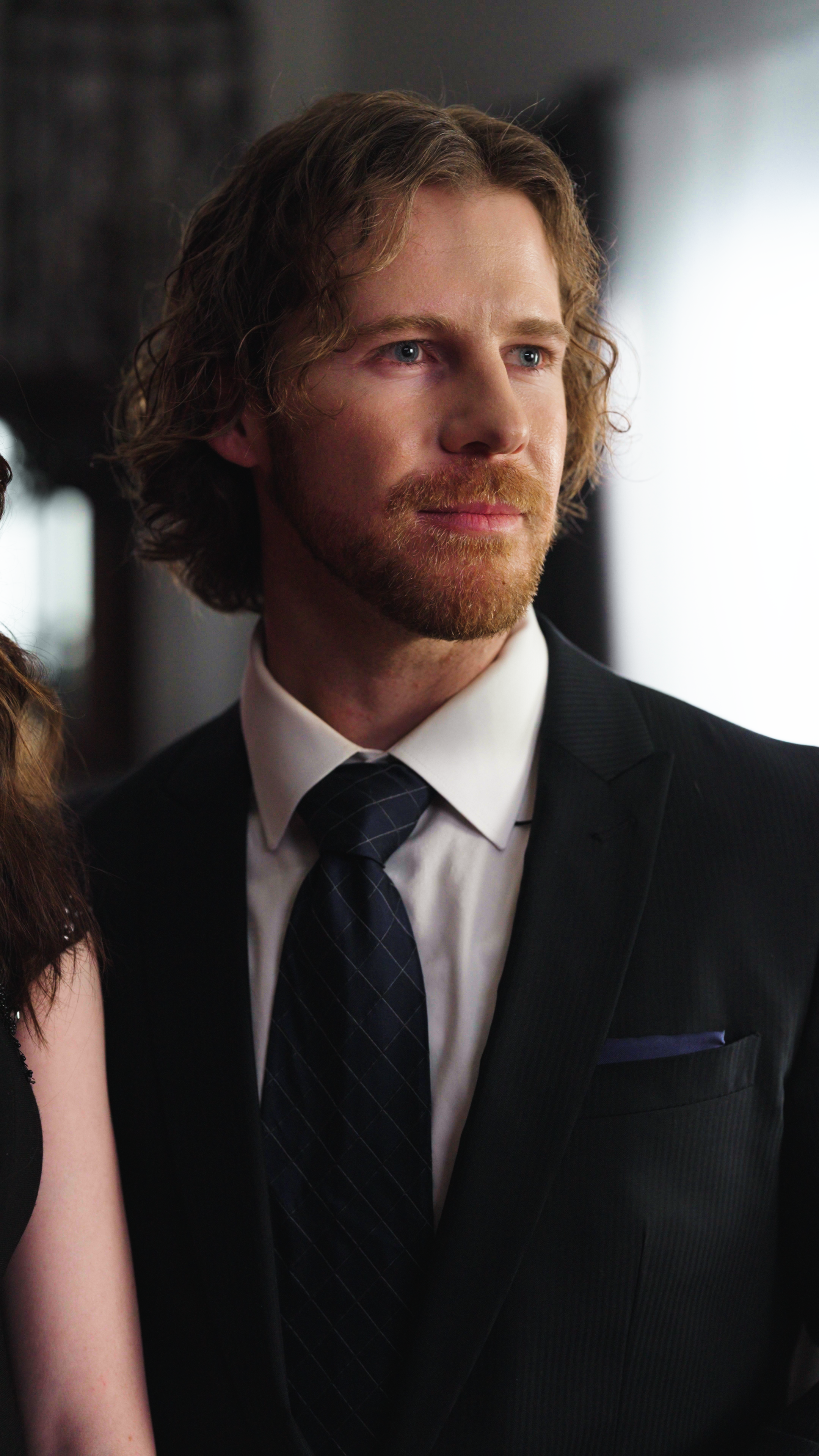 A man with wavy hair and a trimmed beard wears a black suit and dark tie, standing indoors and looking off to the side with a composed but serious expression. Soft natural light falls across his face, emphasizing the tense mood of the moment.