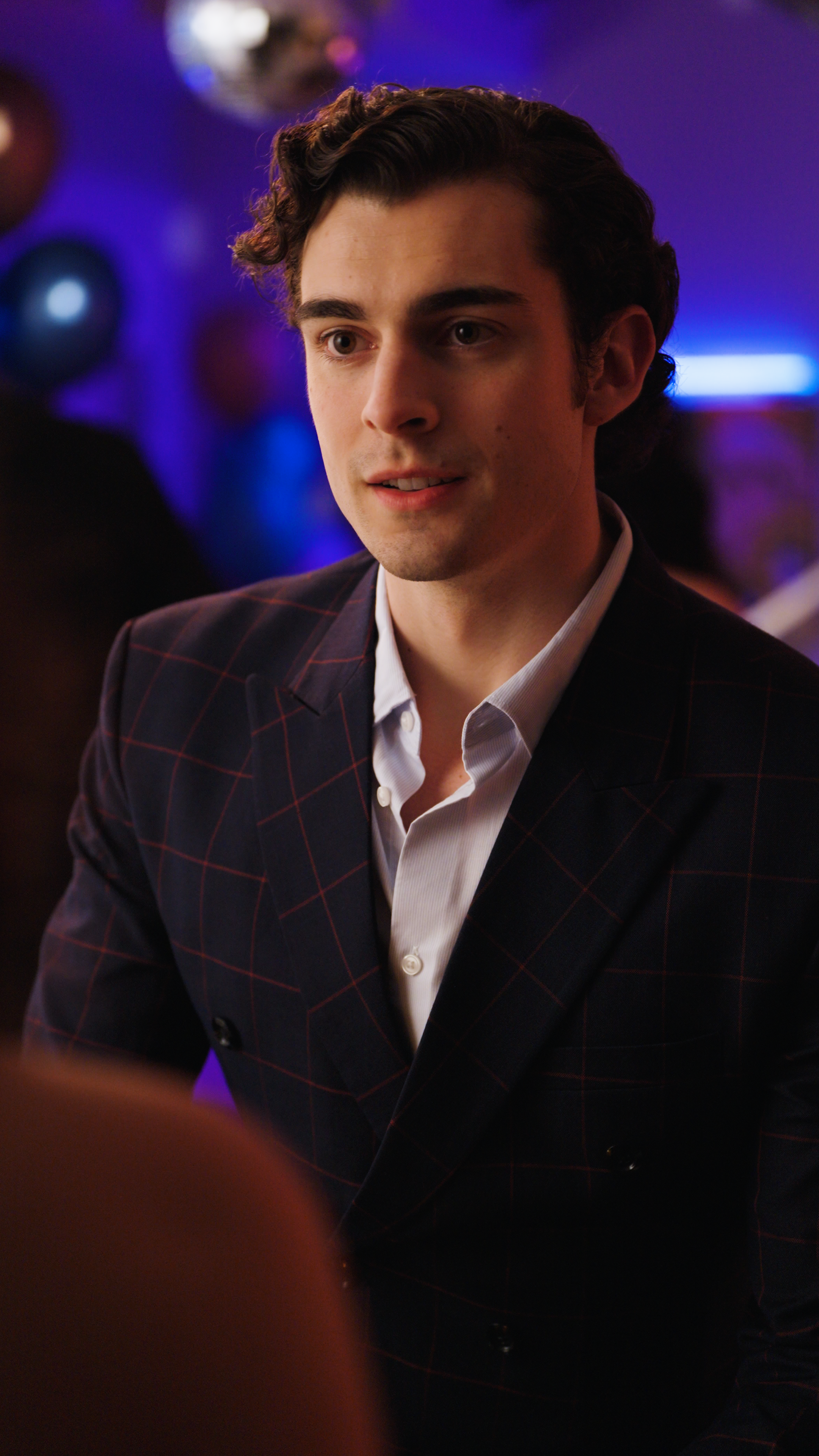 Man in a dark checkered suit jacket speaking to someone at a party with purple and blue lighting.
