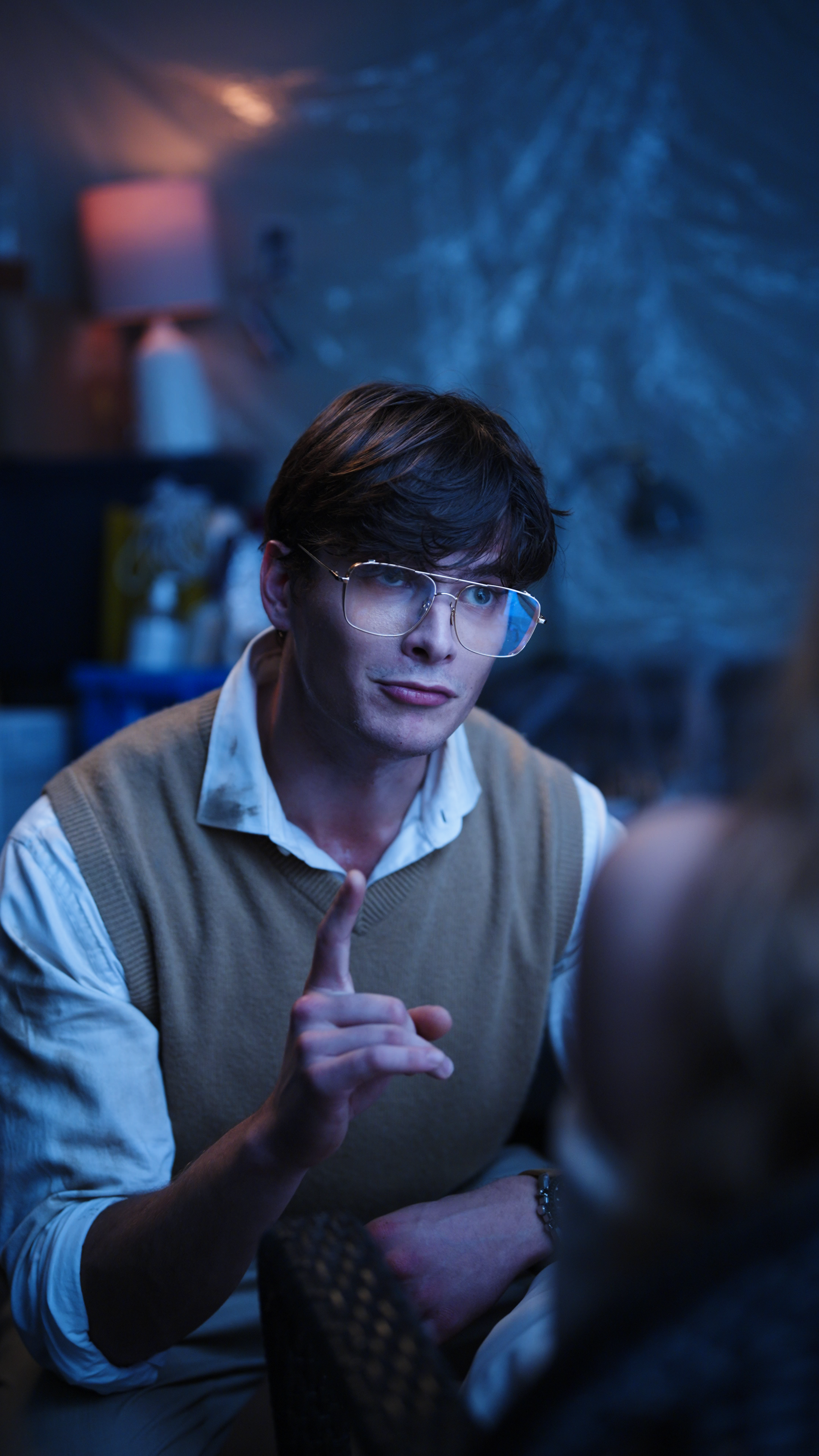 Young man wearing glasses, a white shirt, and a tan sweater vest gesturing with his finger under cool blue moonlight indoors.