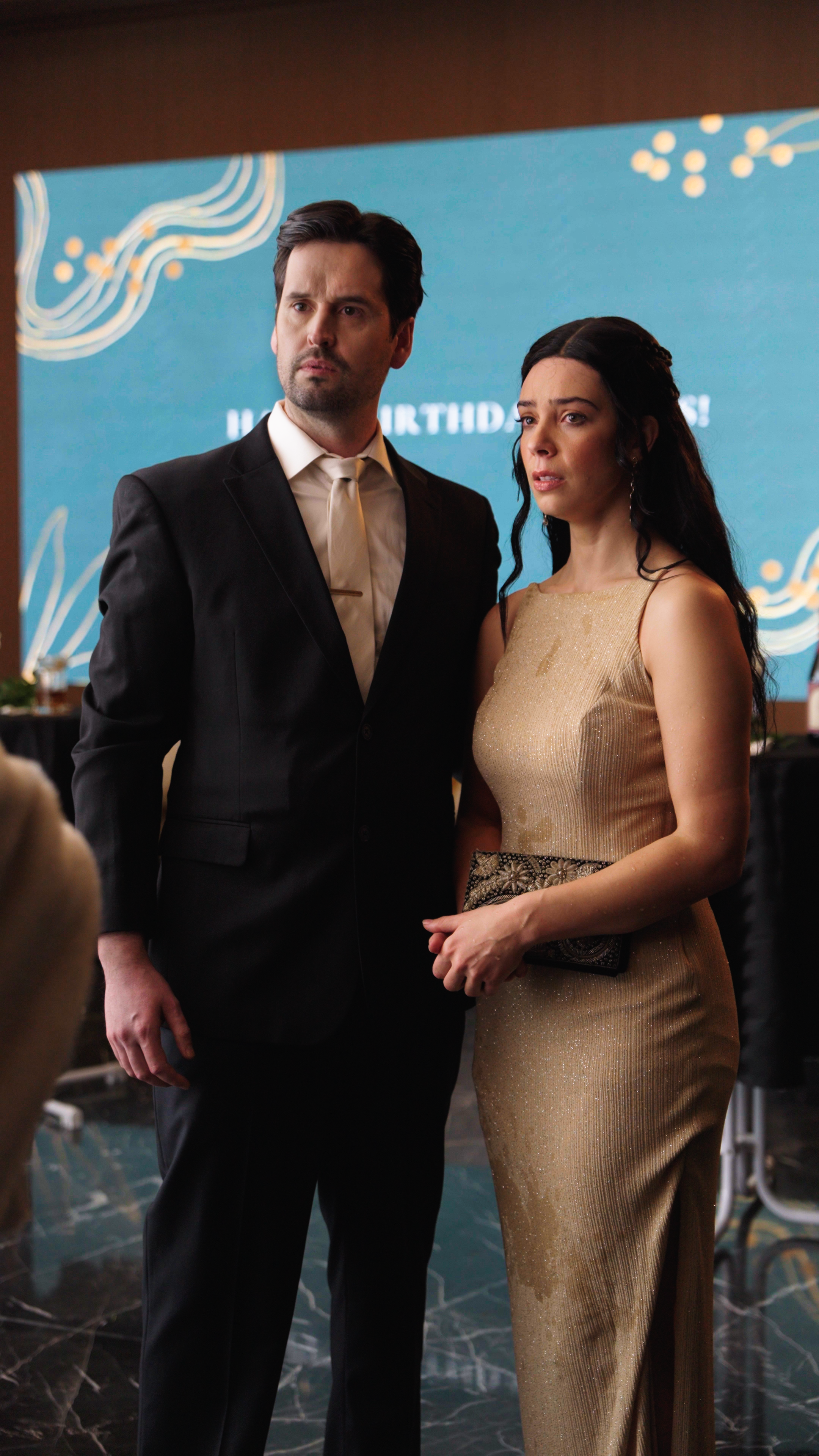 A formally dressed couple stands together at what appears to be a gala or corporate event. The man wears a black suit with a light tie, and the woman wears a fitted gold evening gown, holding a clutch. Both look off to the side with concerned, unsett