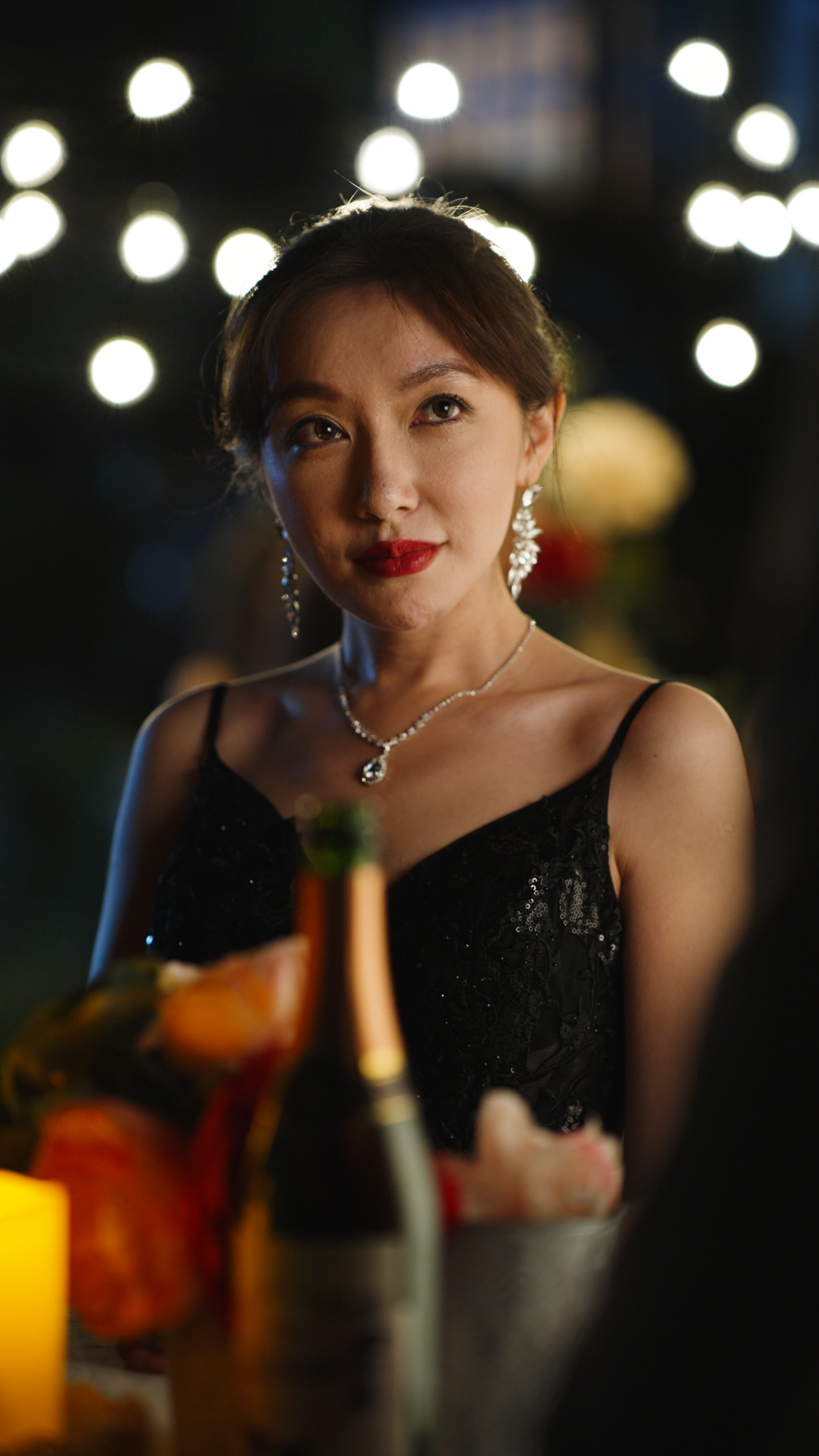 Woman in a black evening dress wearing statement earrings and a pendant necklace seated at a candlelit table with a champagne bottle and flowers in the foreground at an outdoor evening event.