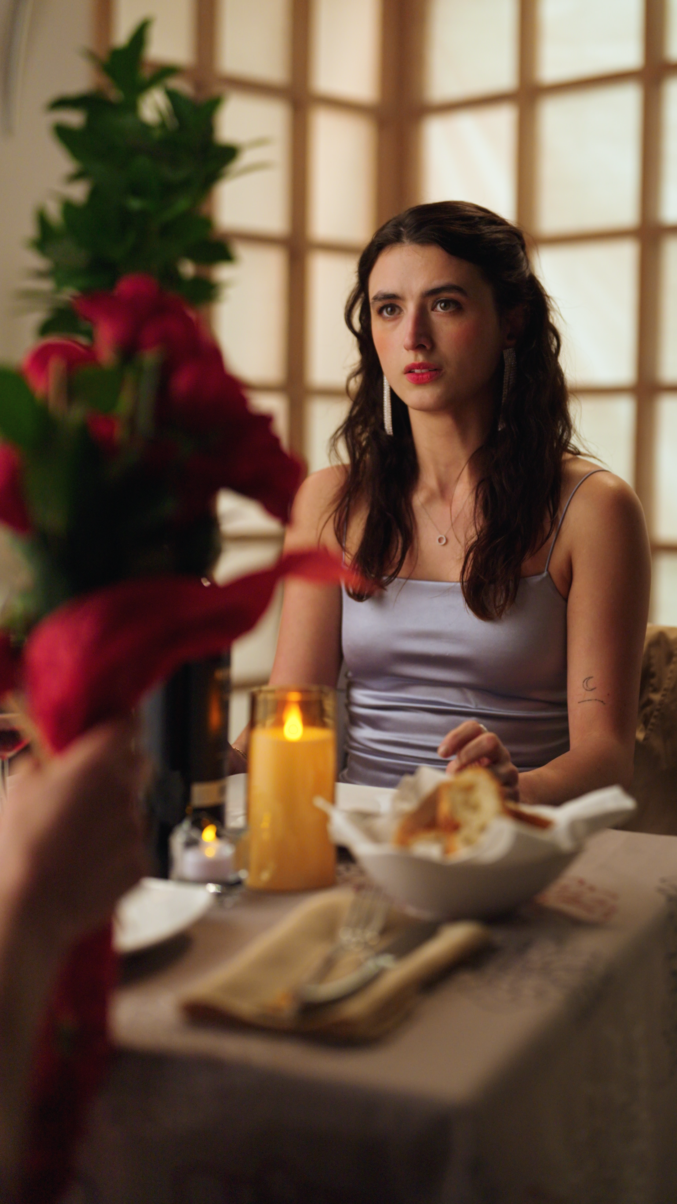 Woman in a lavender satin dress seated at a candlelit dinner table with red flowers and wine bottles in the foreground.