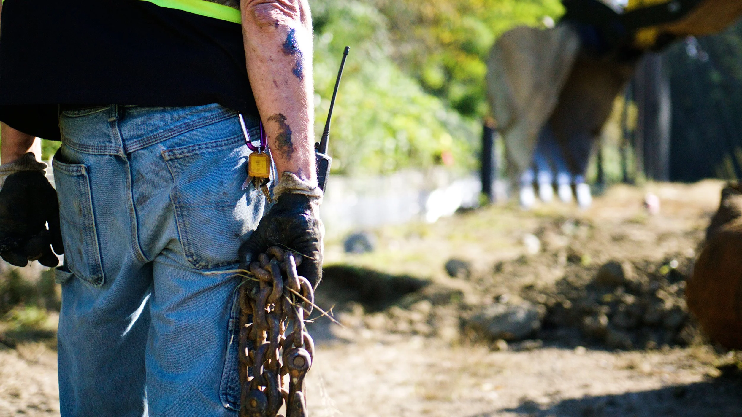 Construction worker holding heavy chain at a demolition site with excavator in the background.