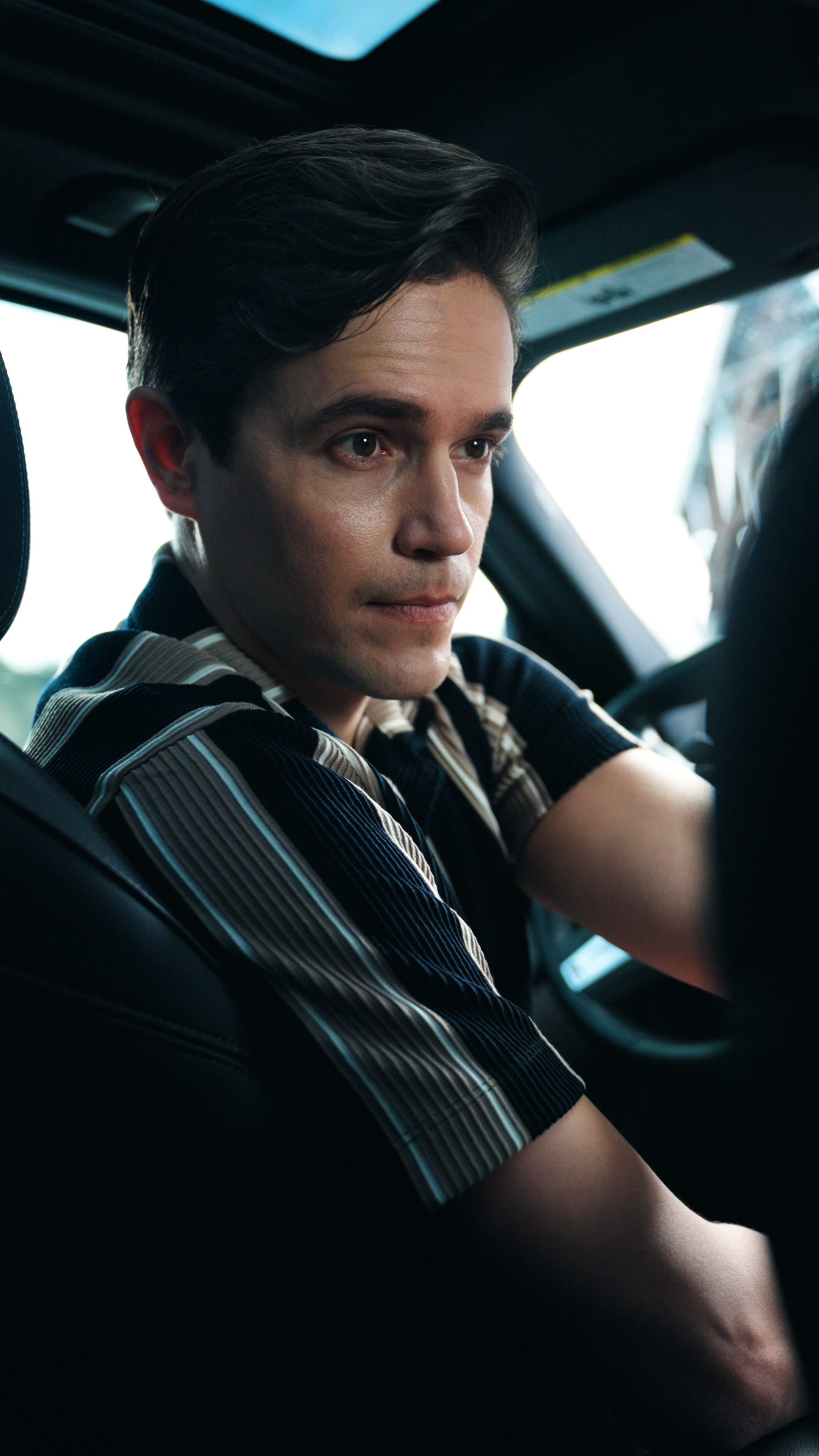 Man in a striped short-sleeve shirt seated in the driver’s seat of a car, looking intently toward a woman in the passenger seat.