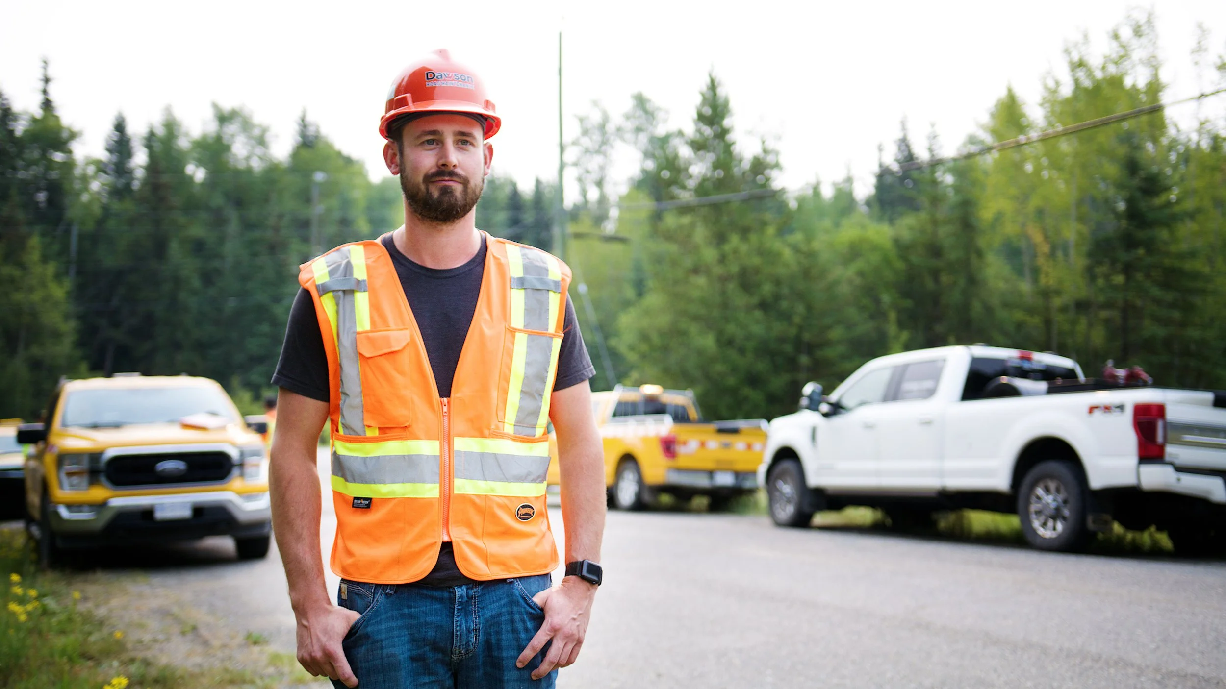 Road construction worker in high-visibility vest and hard hat standing on a rural roadway with service trucks behind him.