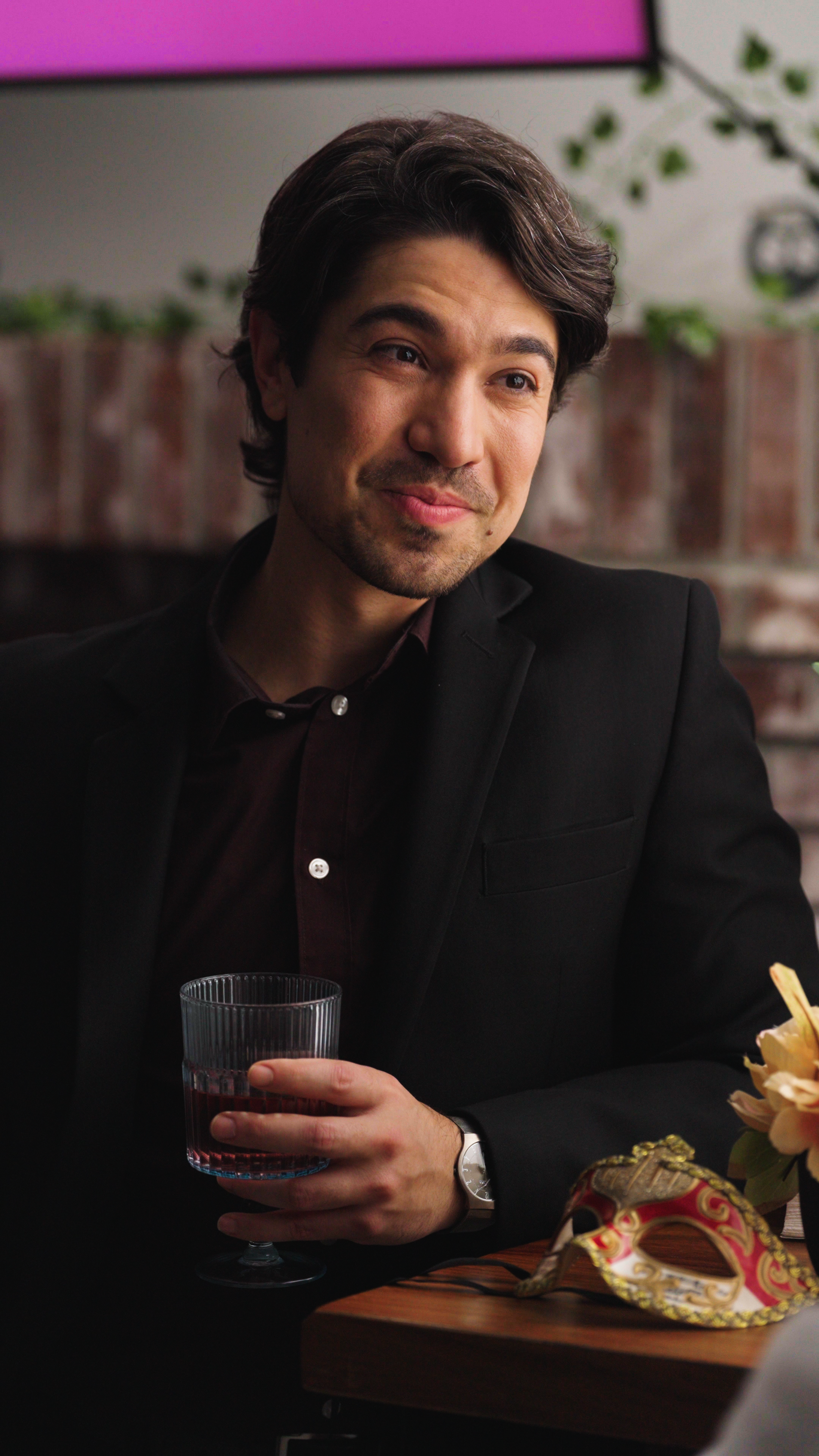 A well-dressed man in a black blazer and dark shirt sits at a table, holding a short glass with a dark red drink. He wears a subtle smile that suggests confidence or amusement. A decorative red-and-gold masquerade mask rests on the table in front of 