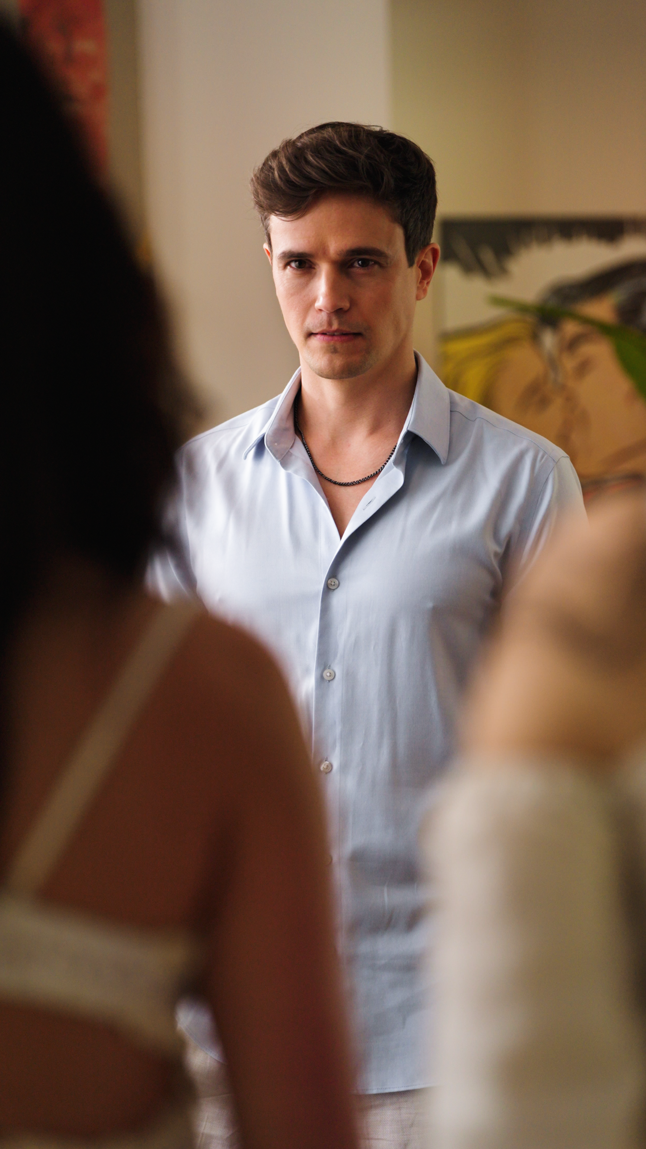 A man in a light blue button-down shirt stands indoors, facing two people in the foreground. His expression is serious and focused, suggesting a tense or important confrontation in a softly lit home setting.