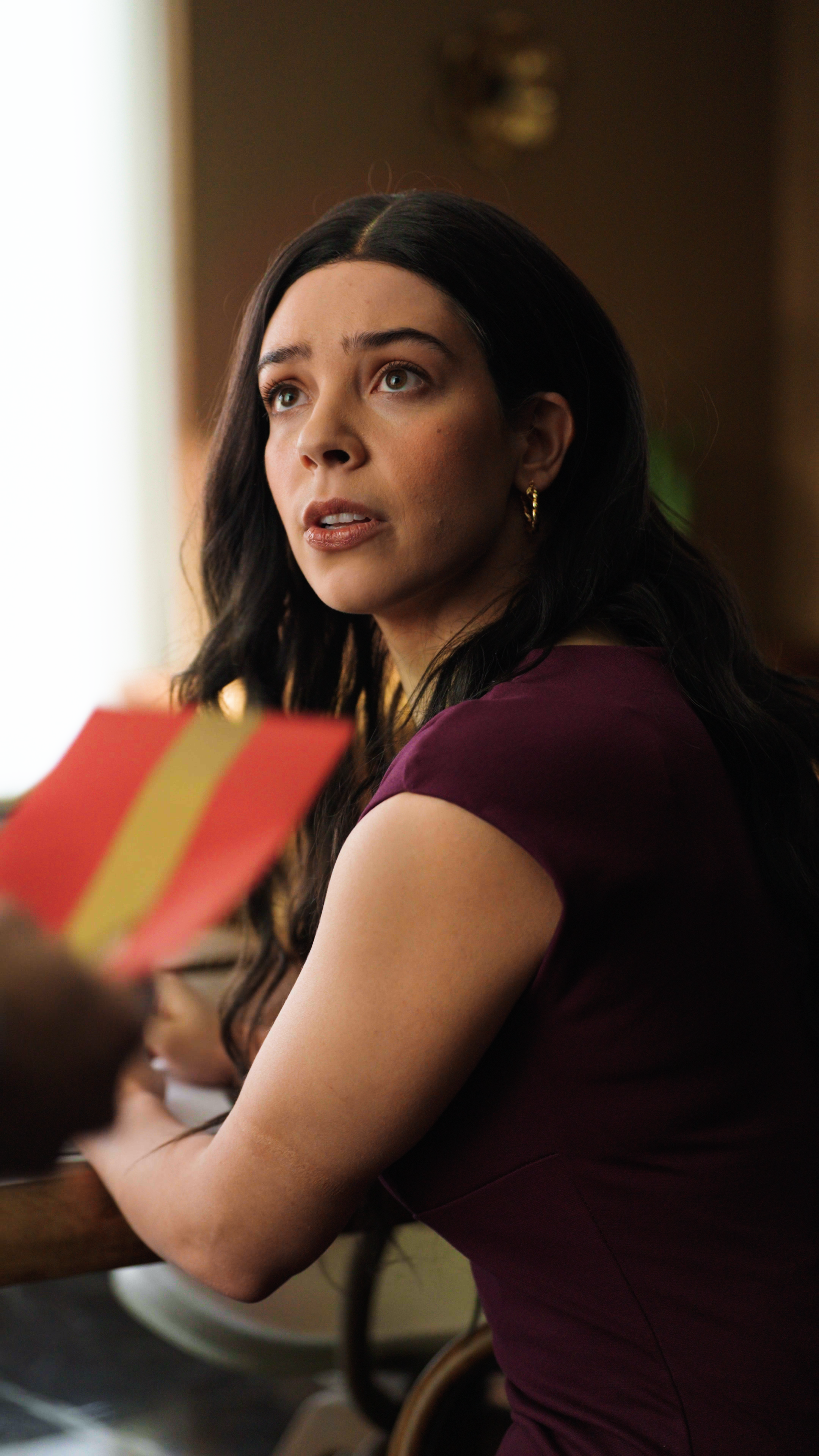 A woman in a burgundy dress sits at a table and turns to look upward with a concerned, slightly startled expression. In the foreground, someone holds a red envelope with a gold stripe toward her. The warm interior lighting and polished wood surfaces 