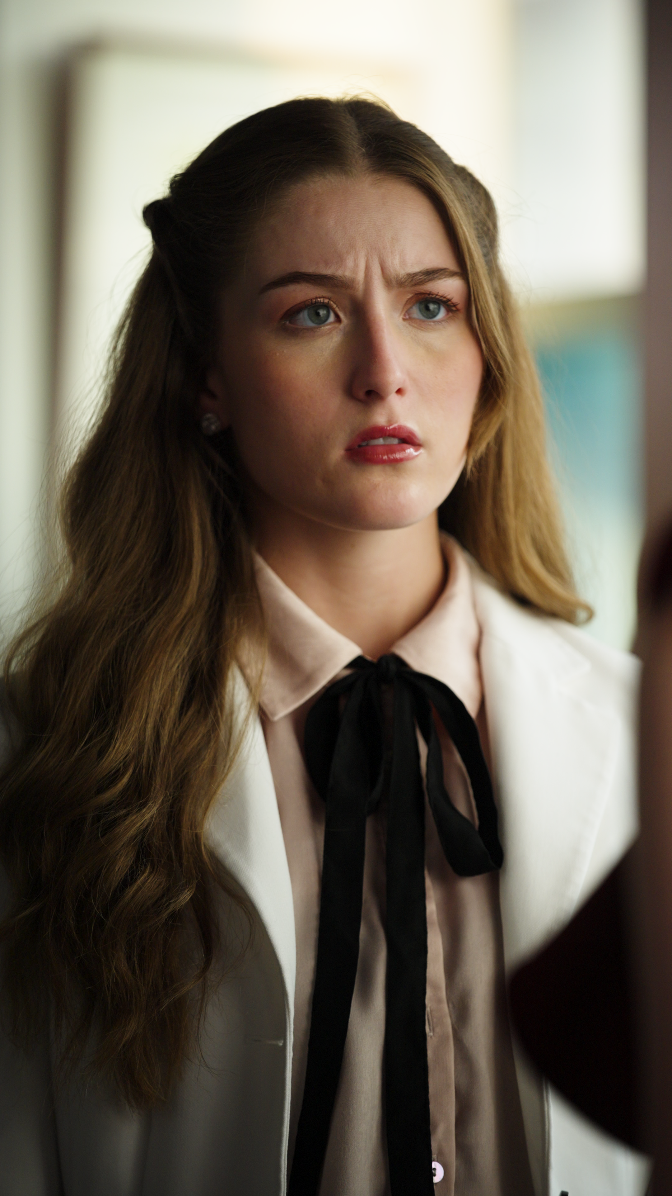 Close-up of a woman in a white blazer and beige blouse with a black bow tie standing indoors.