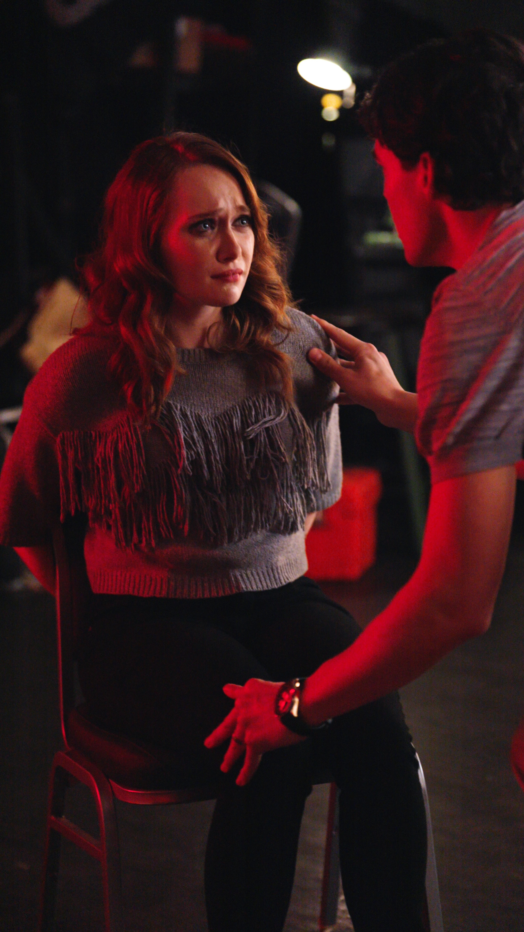 Woman seated on a chair with her hands behind her back while a man places his hand on her shoulder under red lighting indoors.
