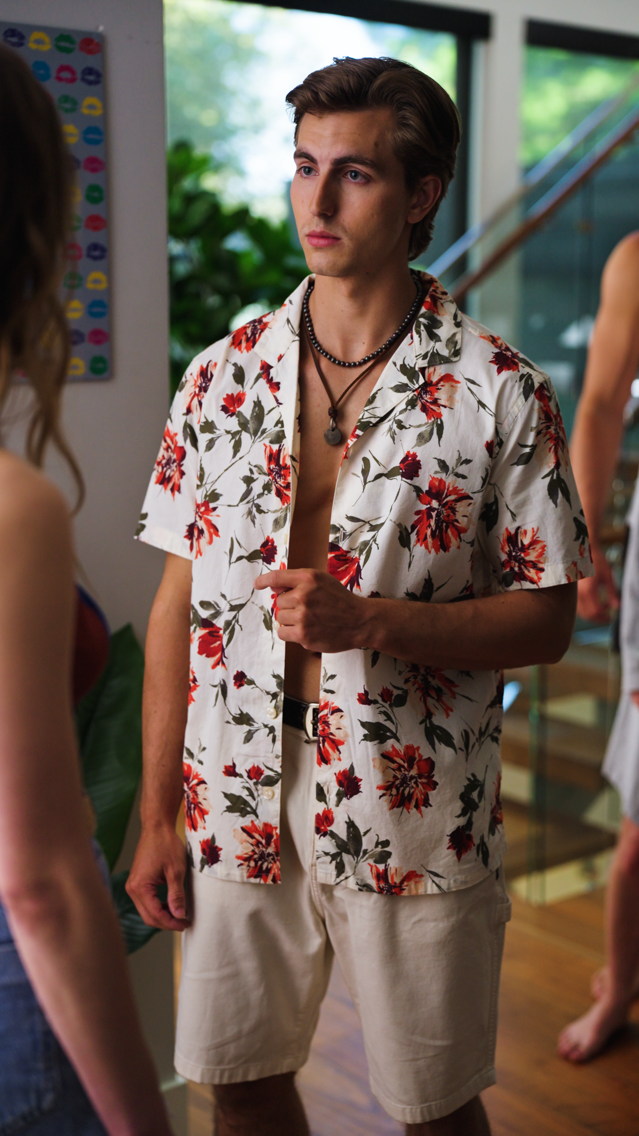 Young man wearing an open floral short-sleeve shirt and light shorts speaking to someone in a modern home interior.