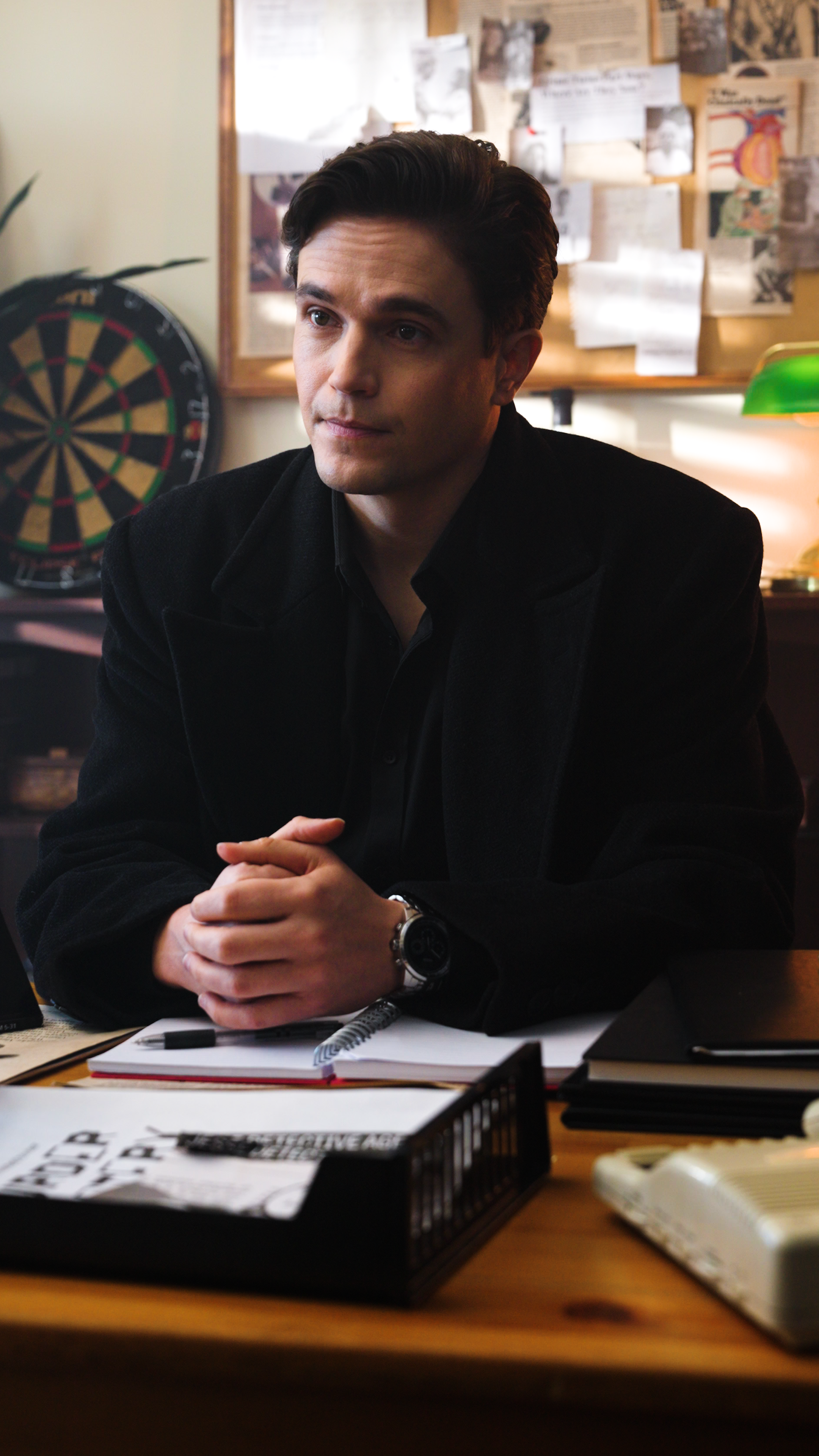 Man in a black jacket seated at a desk with papers and a notebook in front of him, bulletin board and dartboard in the background.