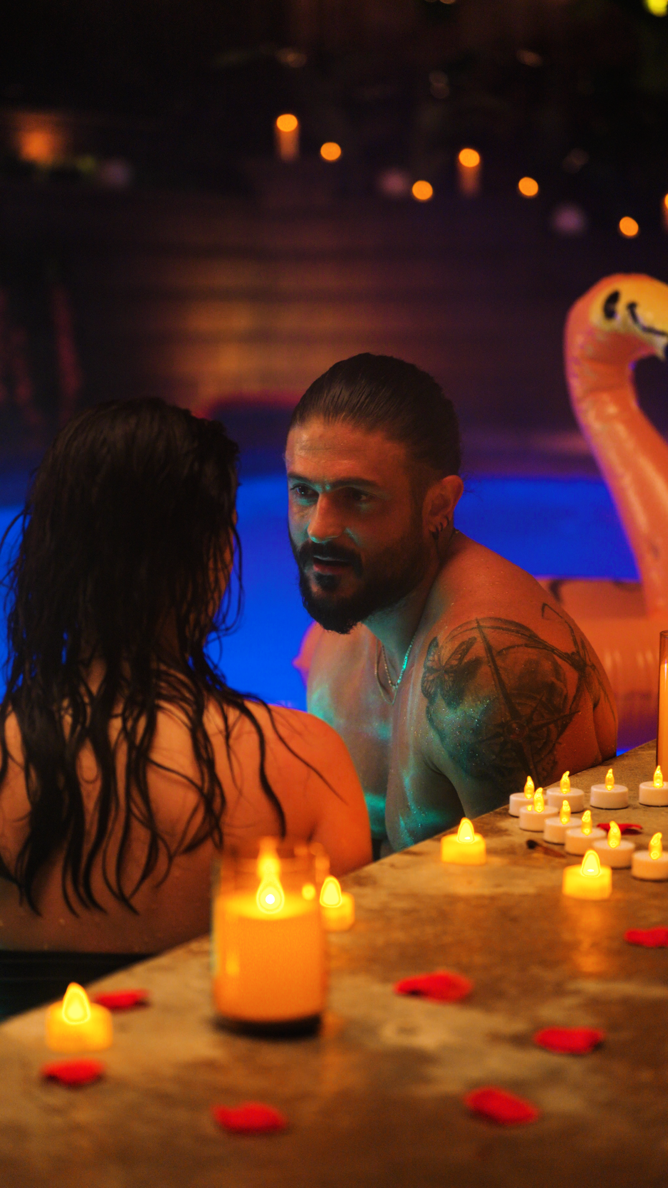 A shirtless man and a woman sitting face to face in a softly lit outdoor pool or hot tub at night. Warm candlelight and scattered rose petals surround them, while cool blue pool lighting creates a contrasting, intimate atmosphere.