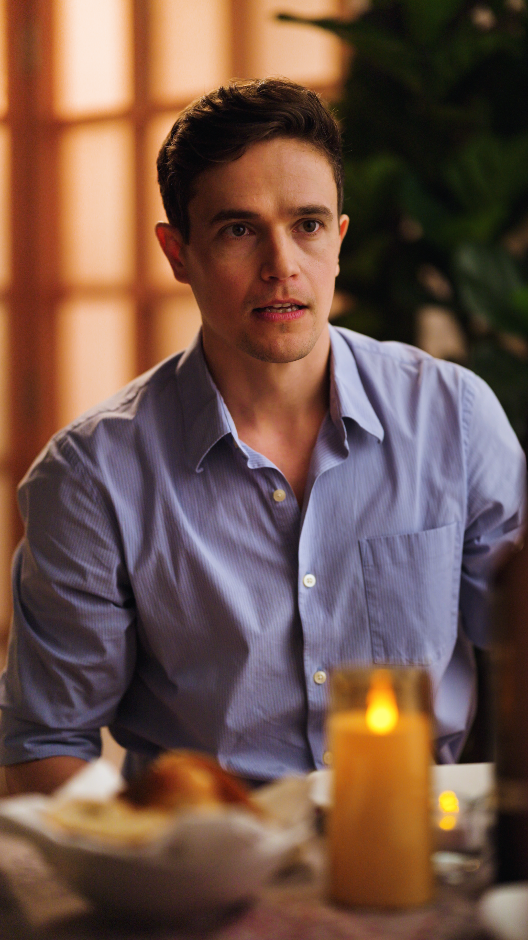 Man in a light blue button-down shirt seated at a dinner table with a lit candle in the foreground.