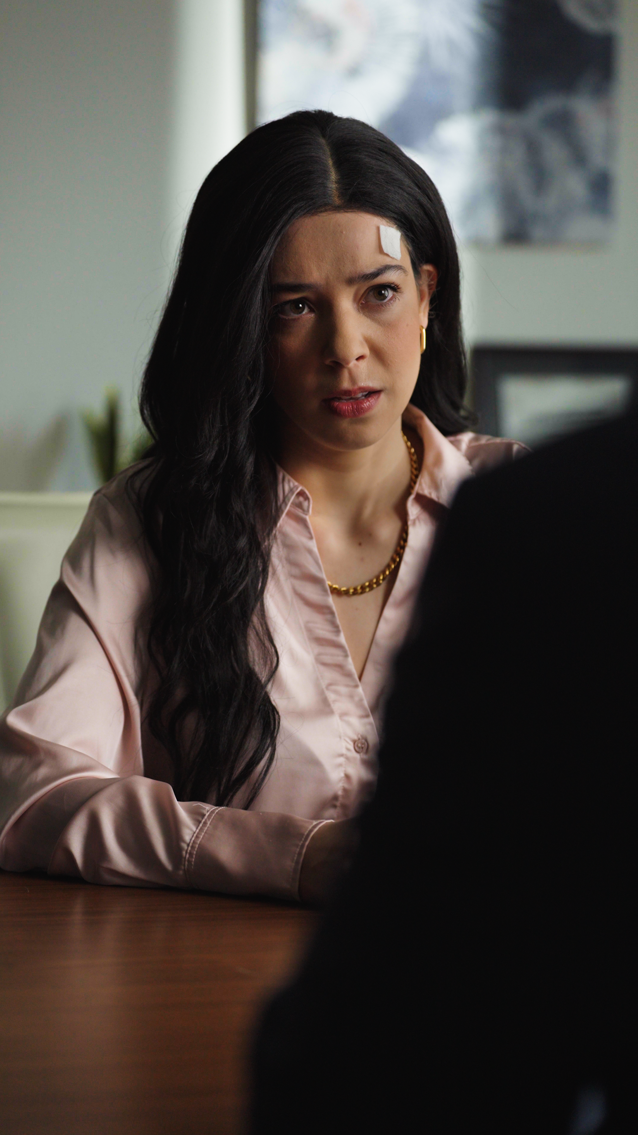 A woman with long dark hair sits at a table indoors, facing someone off camera. She wears a light pink blouse and gold jewelry, and a small bandage is visible on her forehead. Her expression is serious and concerned, suggesting an intense or emotiona