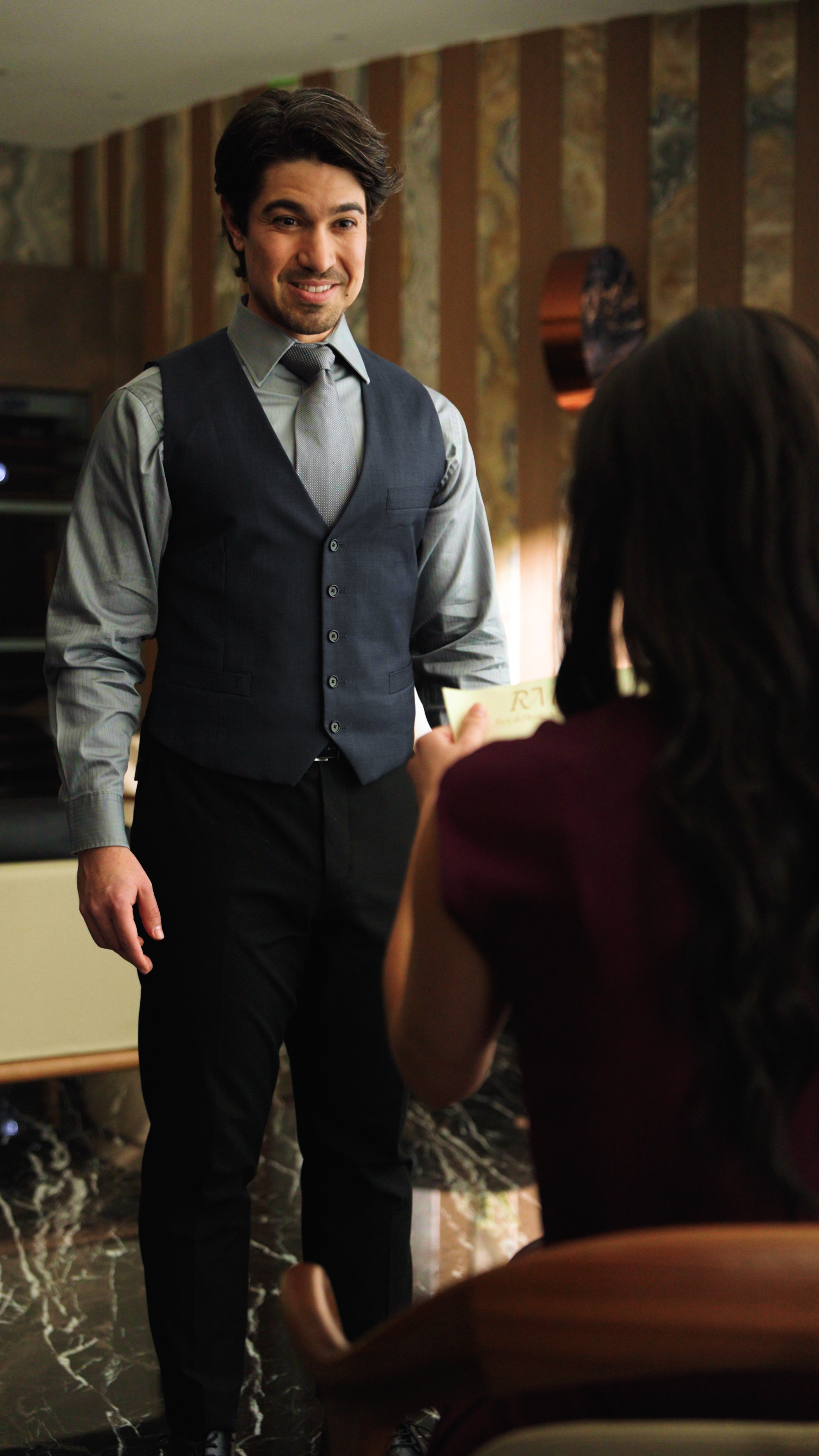 A man wearing a gray shirt, tie, and dark vest stands indoors, smiling slightly as he looks toward a seated woman in the foreground. She appears to be holding a card or envelope toward him. The setting has warm lighting and patterned walls, suggestin