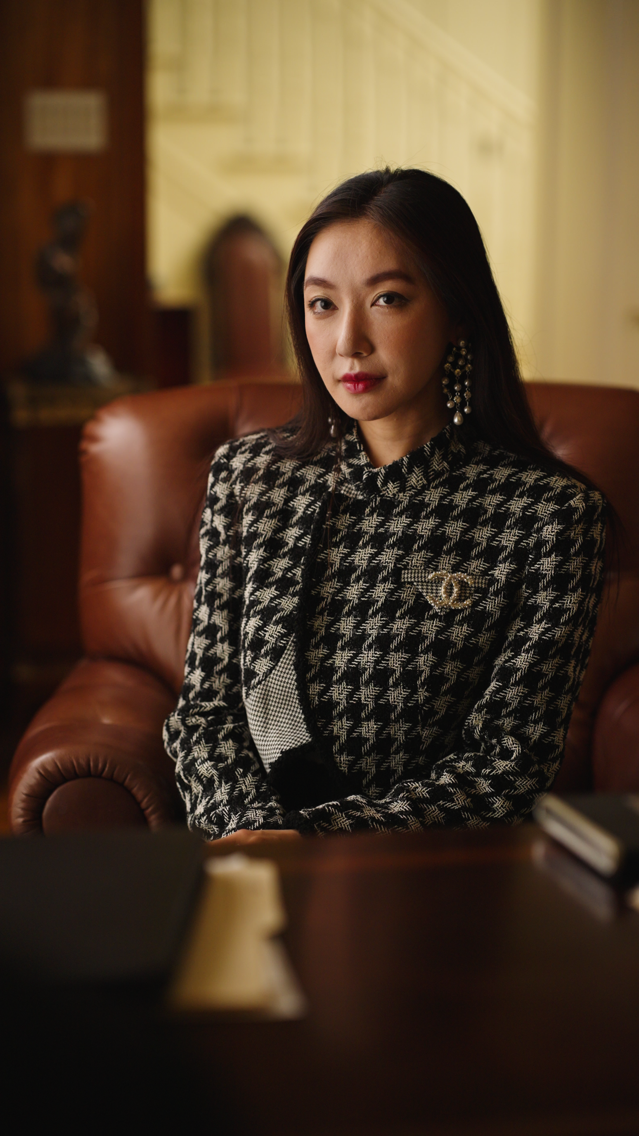 Woman seated in a leather armchair wearing a black and white patterned jacket and statement earrings in an interior setting.