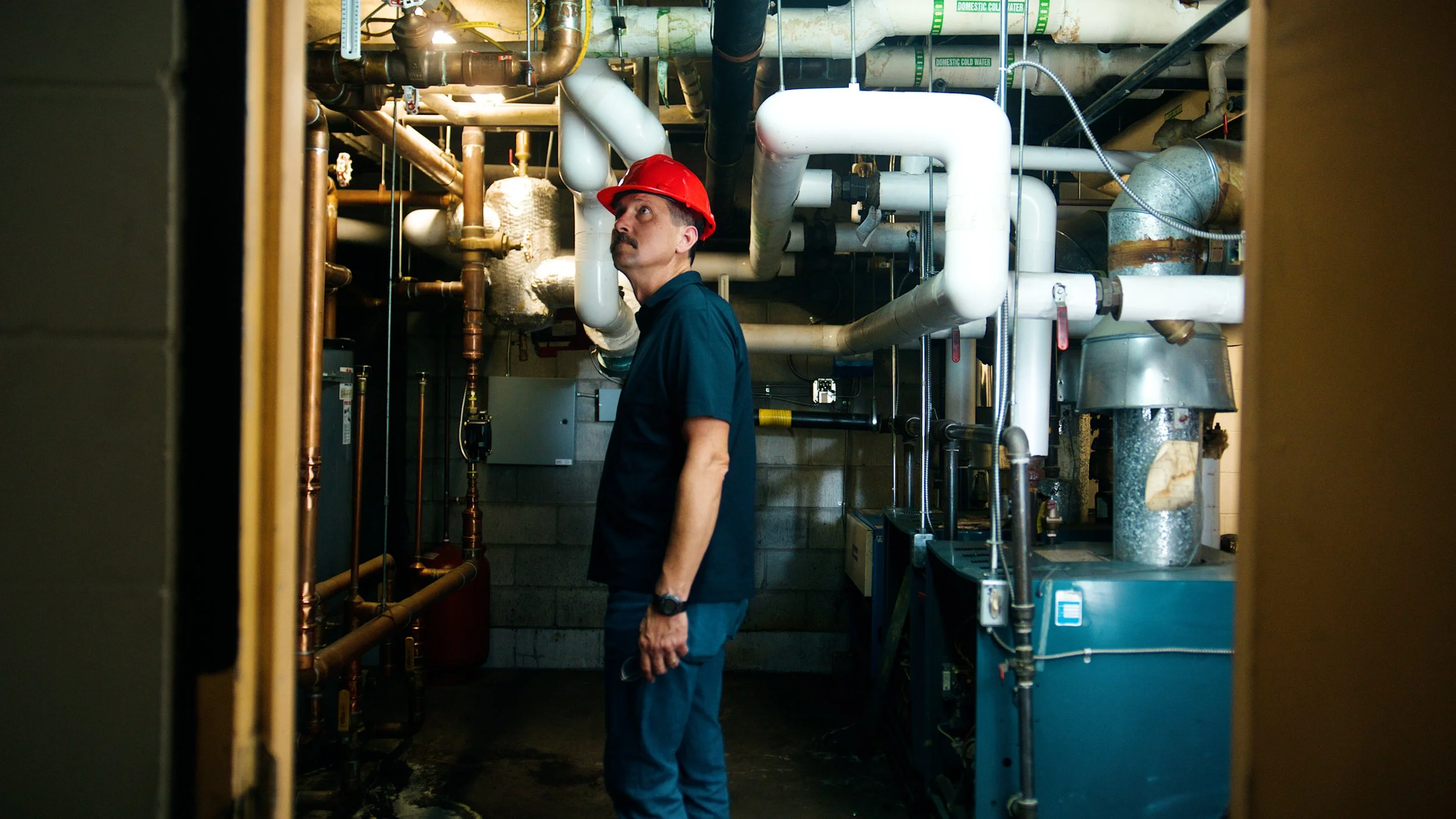 Technician wearing a hard hat inspecting mechanical equipment in a boiler room.