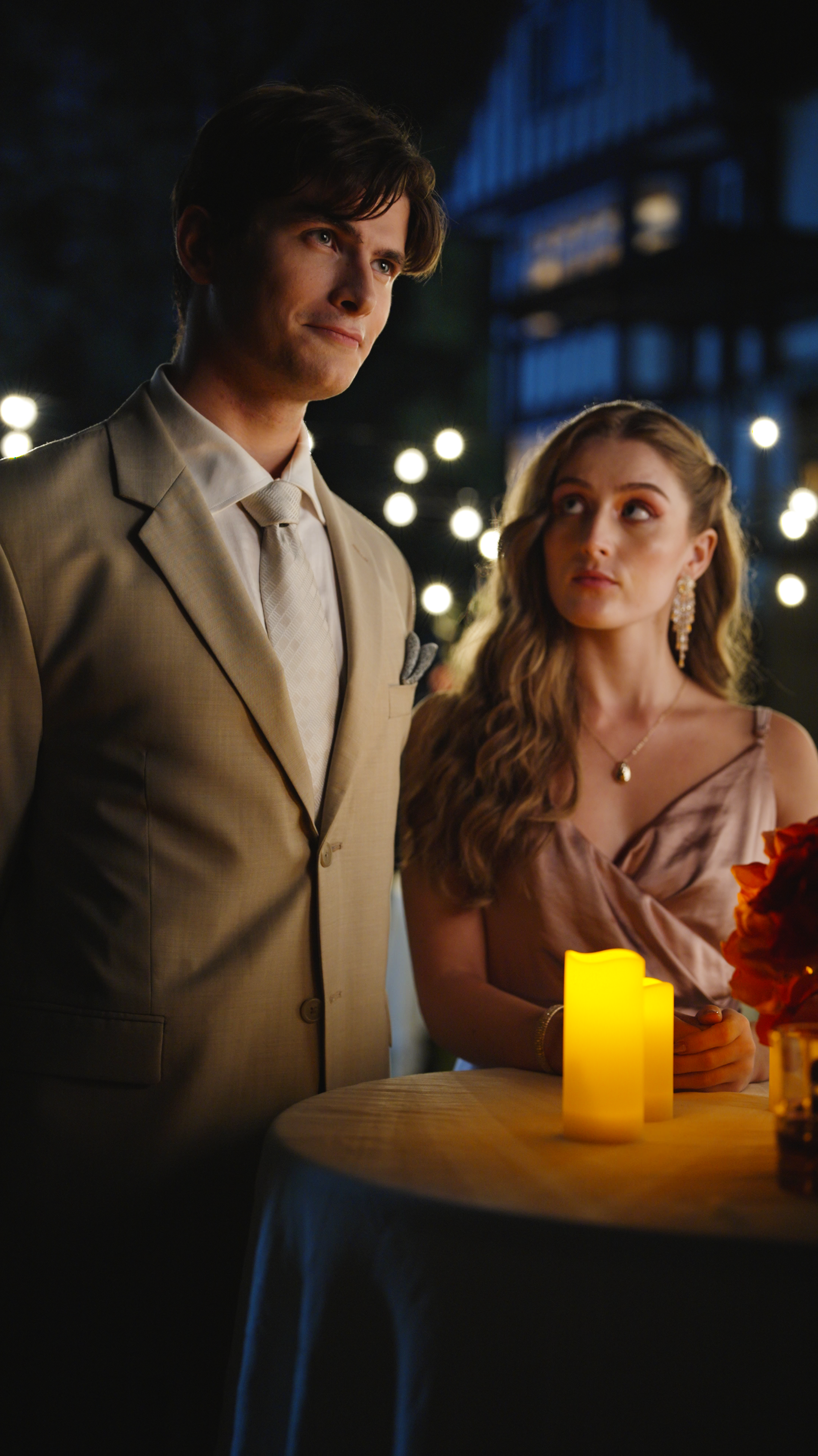Young man in a beige suit and tie standing beside a woman in a satin dress at a candlelit table with string lights in the background at an evening event.