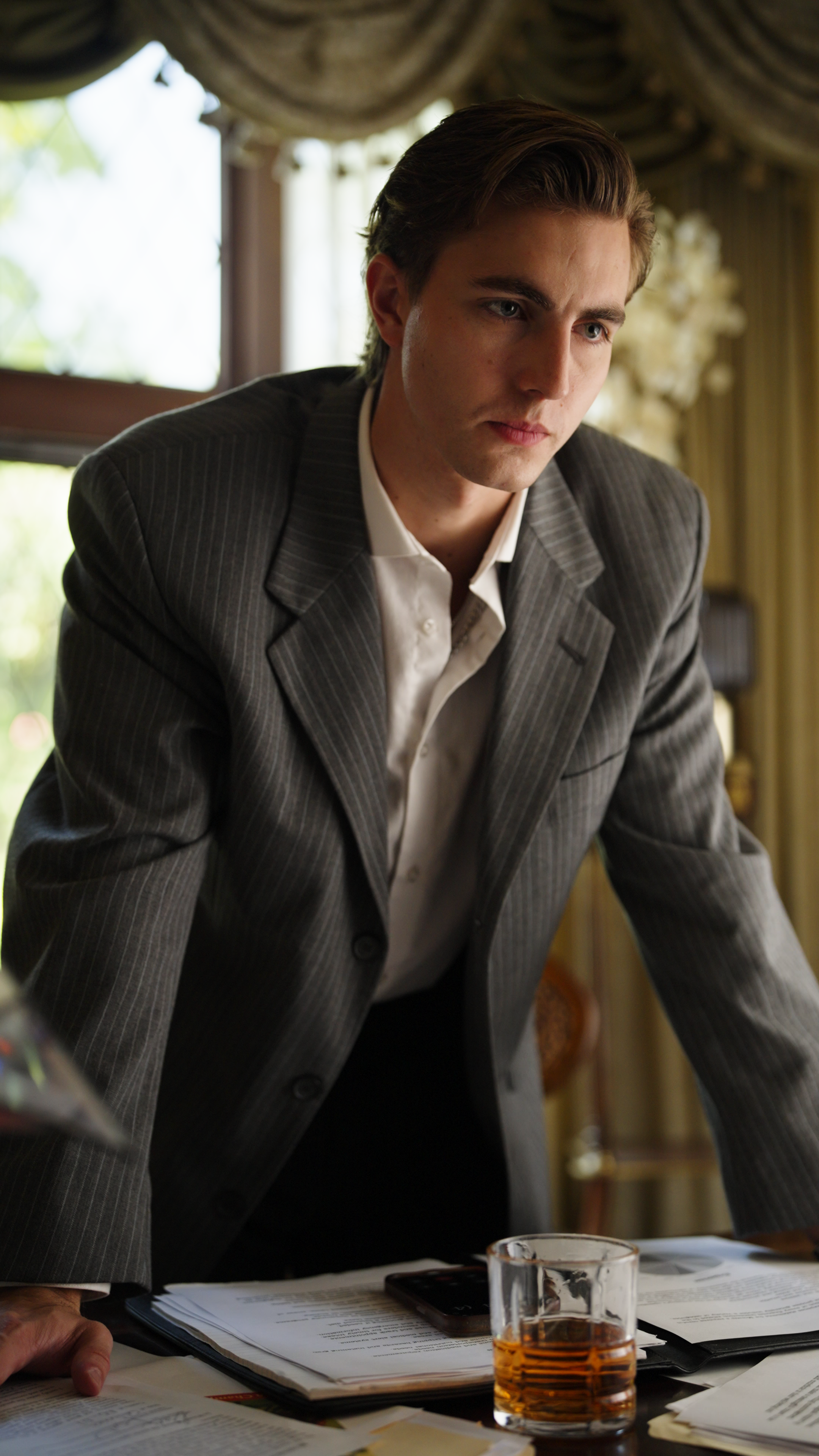 Young man in a pinstripe suit leaning over a desk with documents and a glass of whiskey in an interior setting.