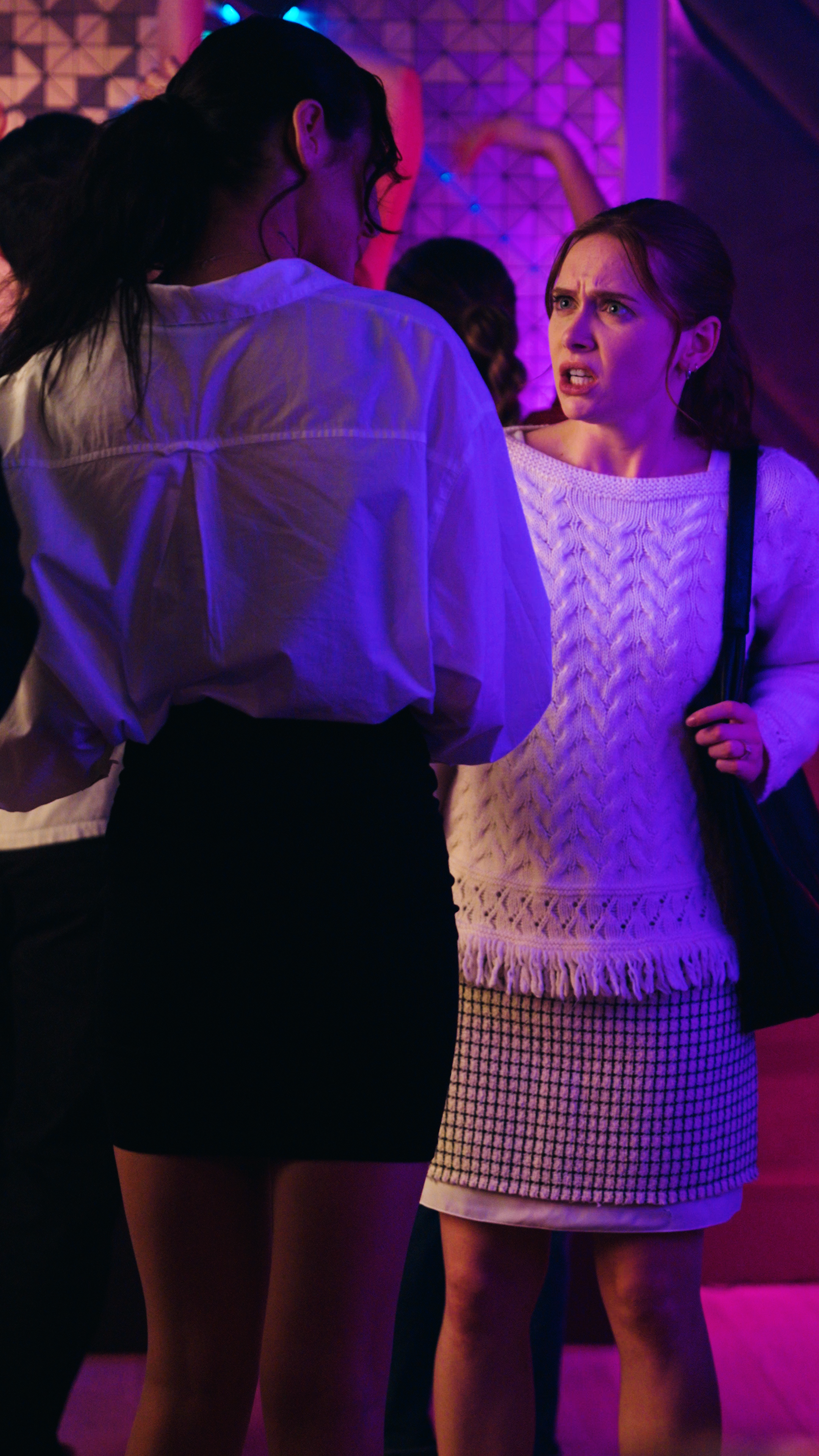Woman in a white sweater confronting another woman in a white blouse and black skirt under purple and pink club lighting.