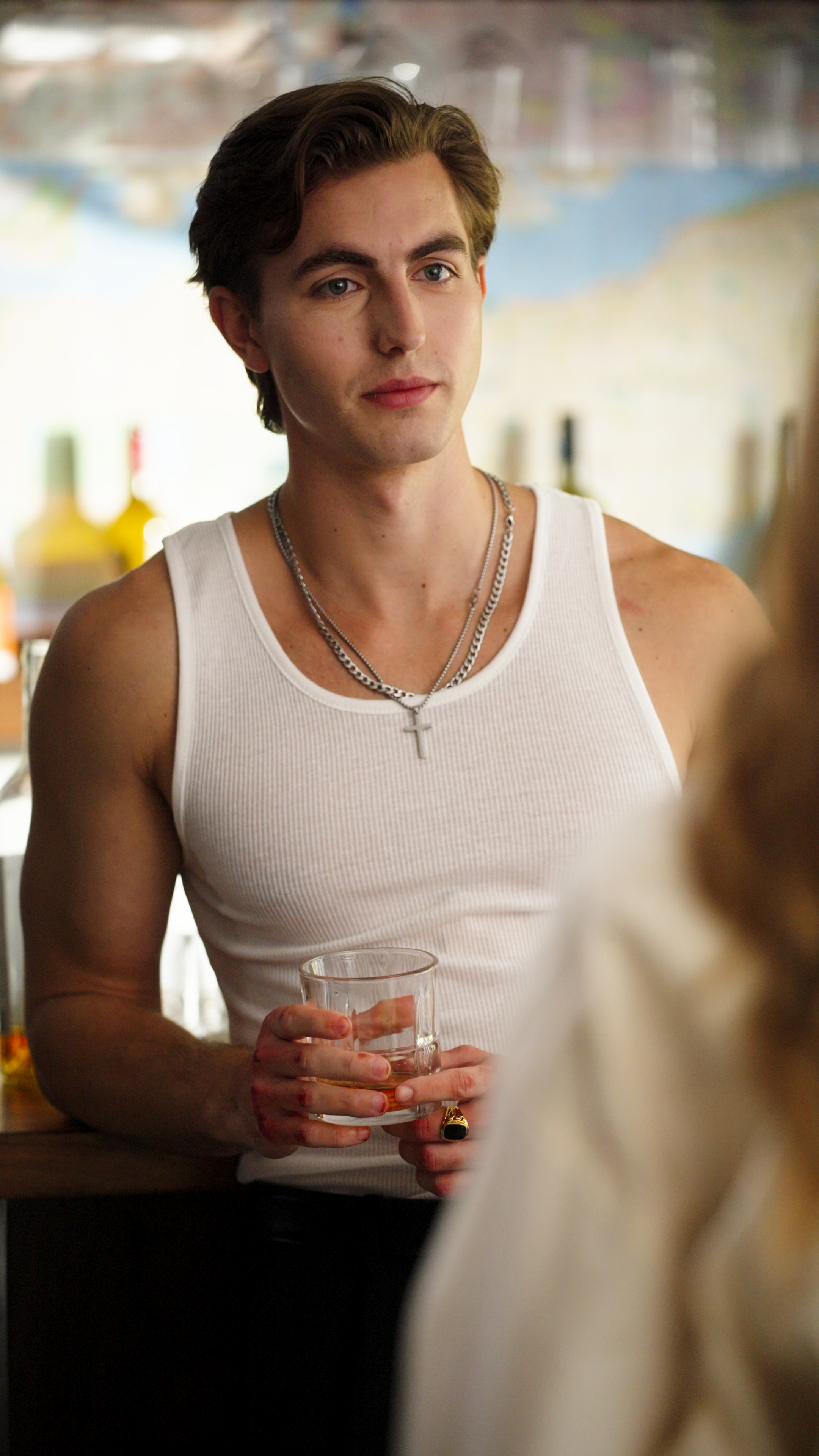 Young man at a bar holding a whiskey glass, wearing a white tank top and silver chain necklace.