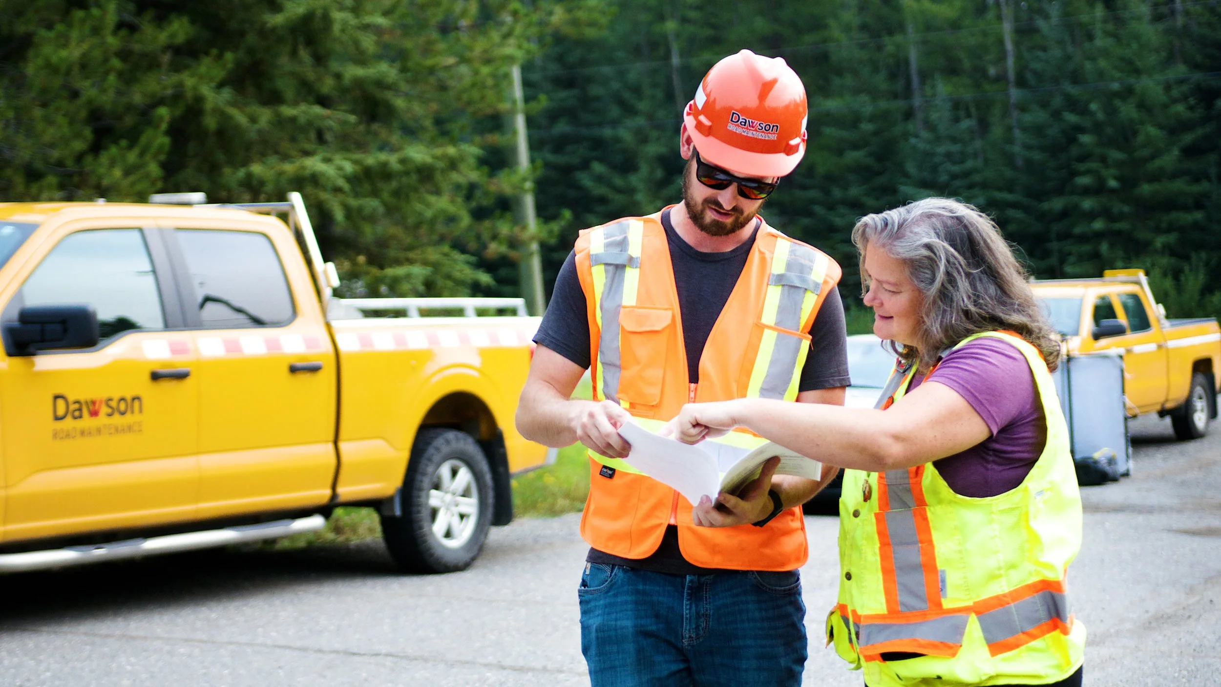 Two road maintenance workers reviewing paperwork in high-visibility safety gear.