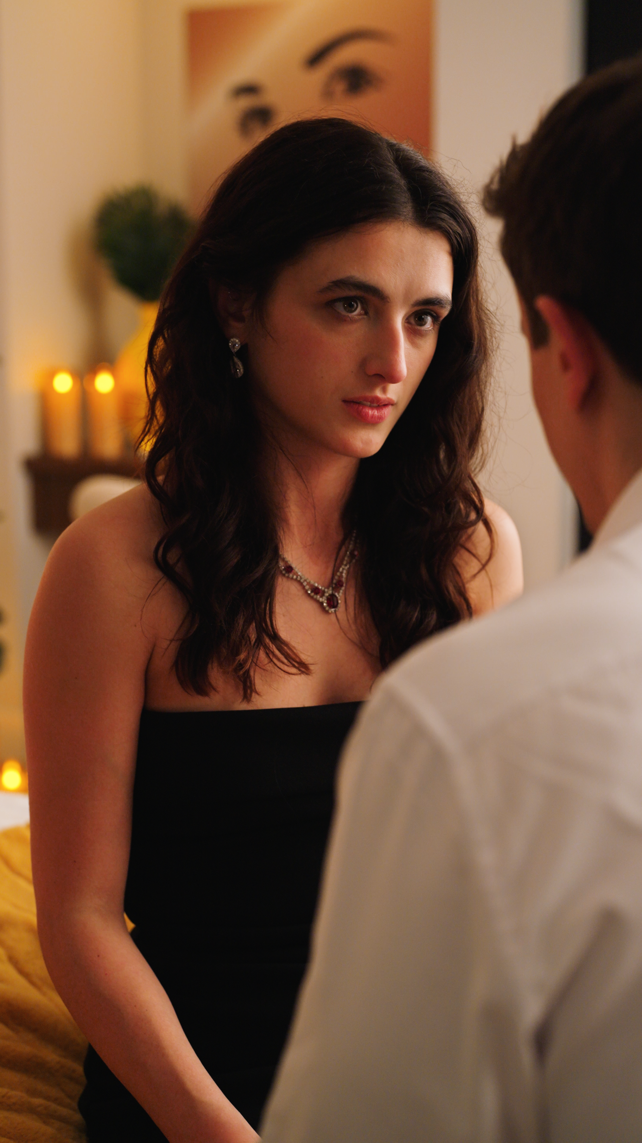 Woman in a black strapless dress looking seriously at a man during a candlelit indoor conversation.