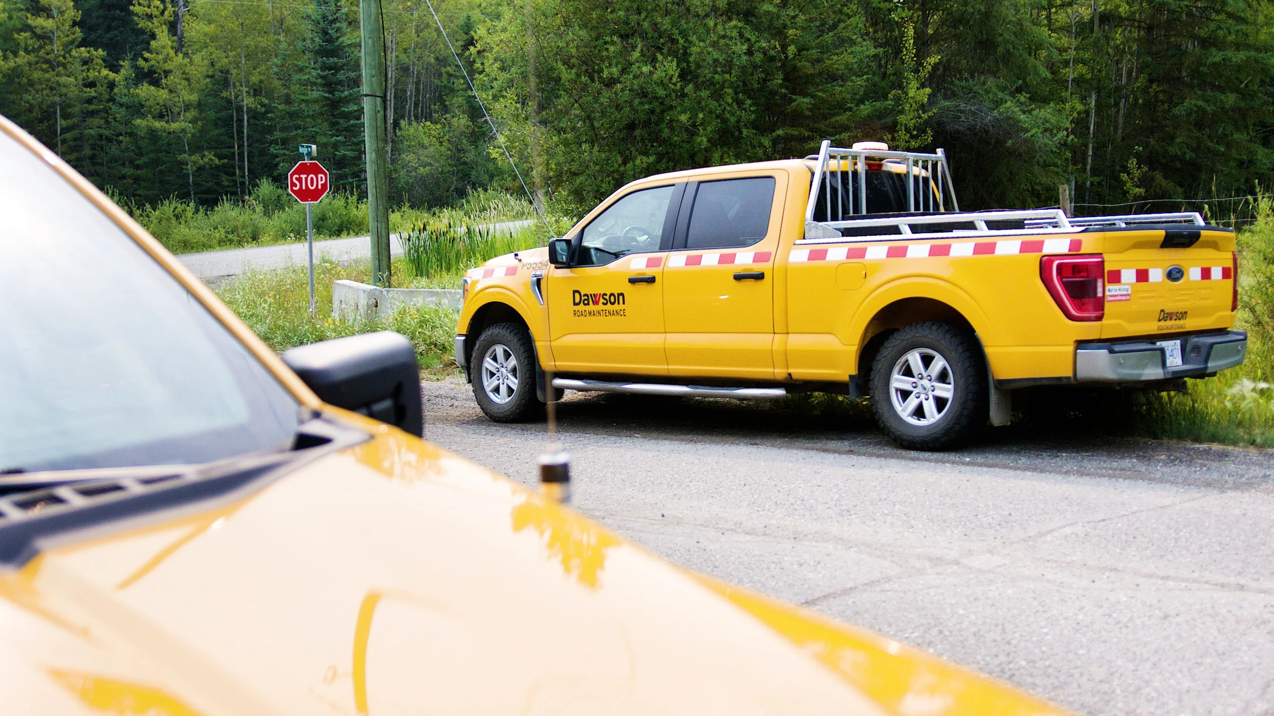 Road maintenance truck parked at a rural intersection with a stop sign and forest backdrop.