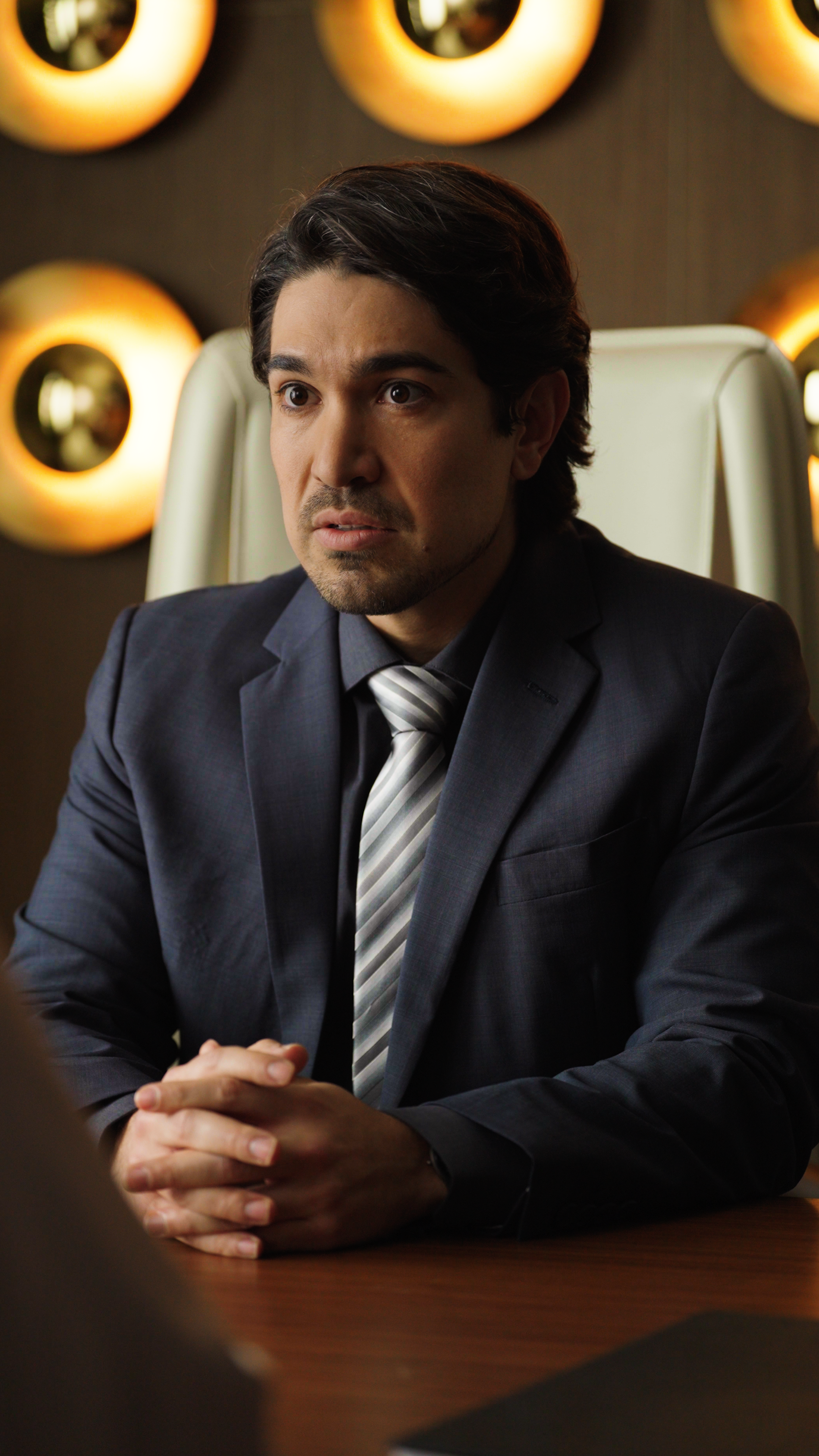 A man in a dark suit and striped tie sits at a desk in a modern office setting. He clasps his hands together and looks forward with a tense, uneasy expression. Circular warm wall lights glow behind him, adding a dramatic backdrop to the serious conve