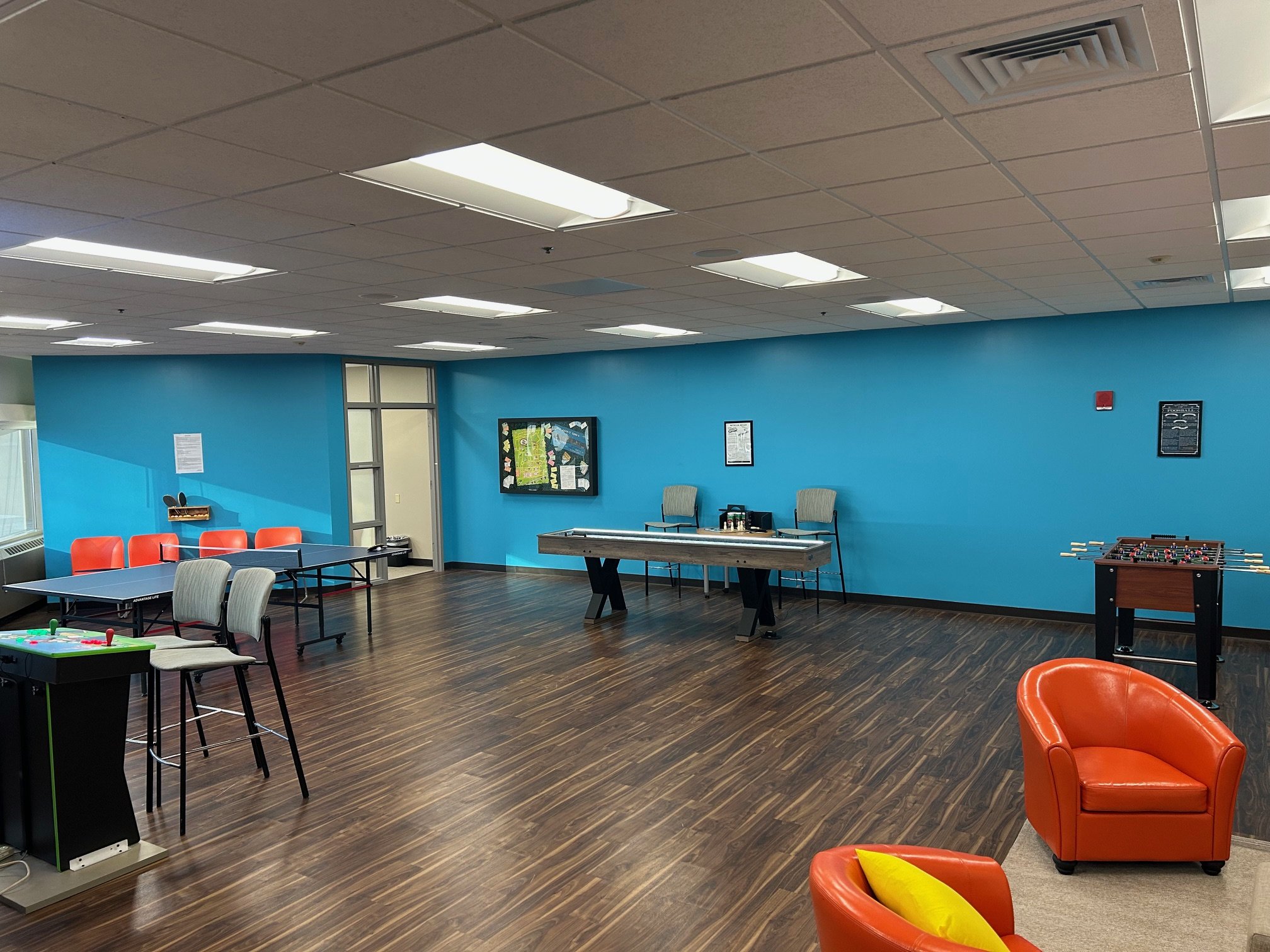 A modern recreation room with a ping pong table, foosball table, orange chairs, and a blue accent wall.
