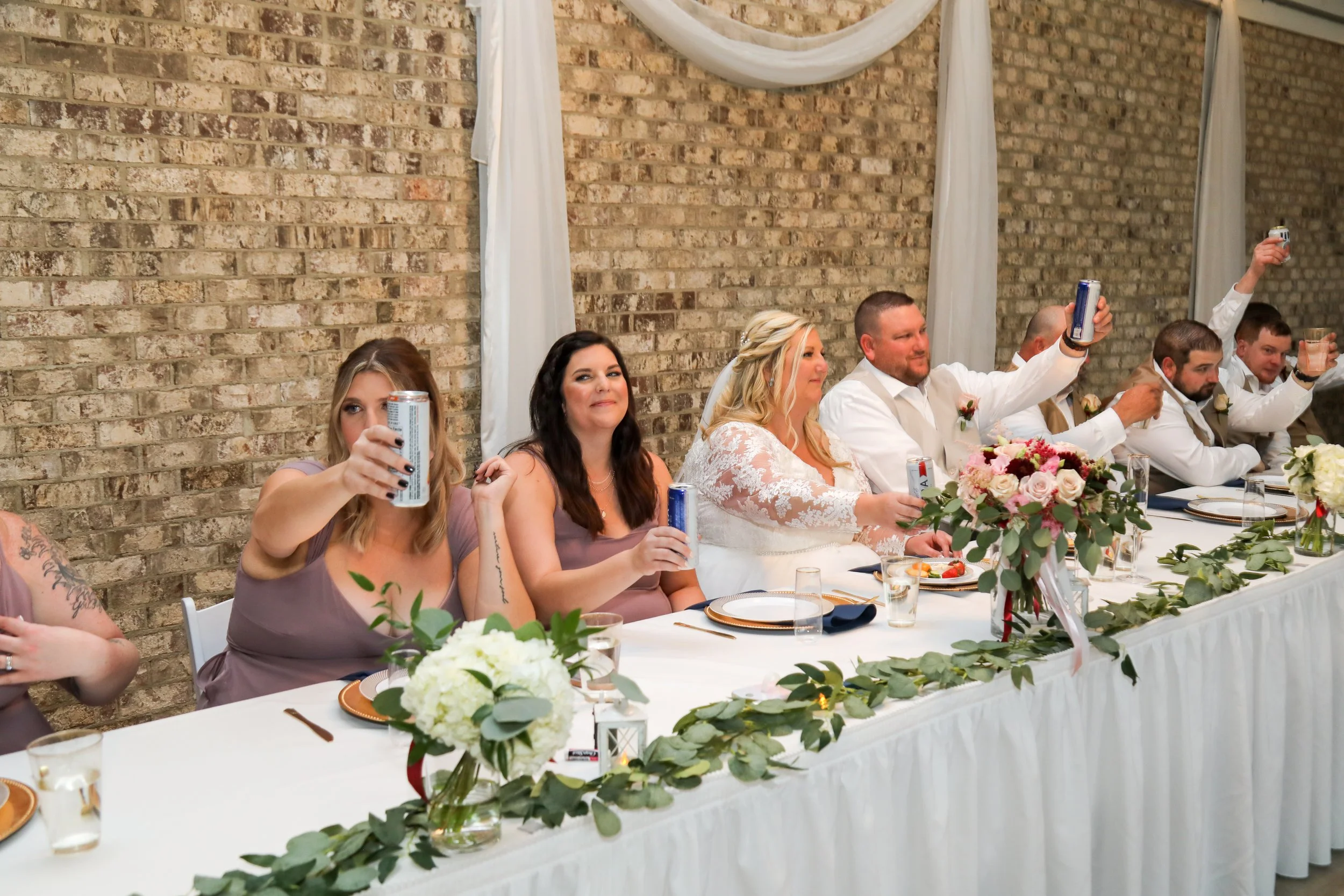 This is a scenic boho rustic view of the head table inside of The Magnolia Barn award-winning reception hall outdoor barn wedding venue featuring an exposed brick wall with natural light in Miamisburg at Magnolia Estate. Surrounded by the Greater Day
