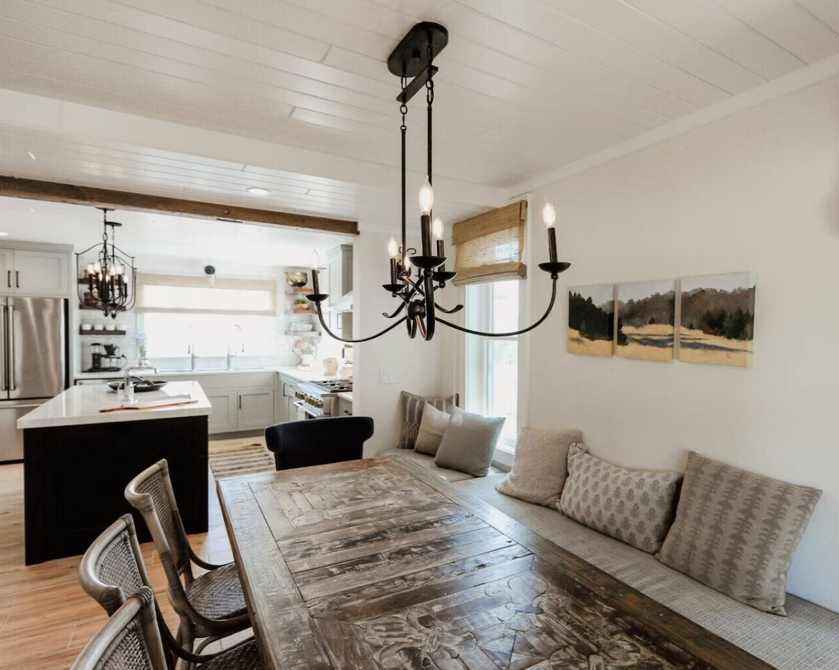 A rustic dining room and kitchen with a chandelier hanging above a wooden dining table. The kitchen has white cabinets, a stainless steel refrigerator, and large windows letting in natural light.