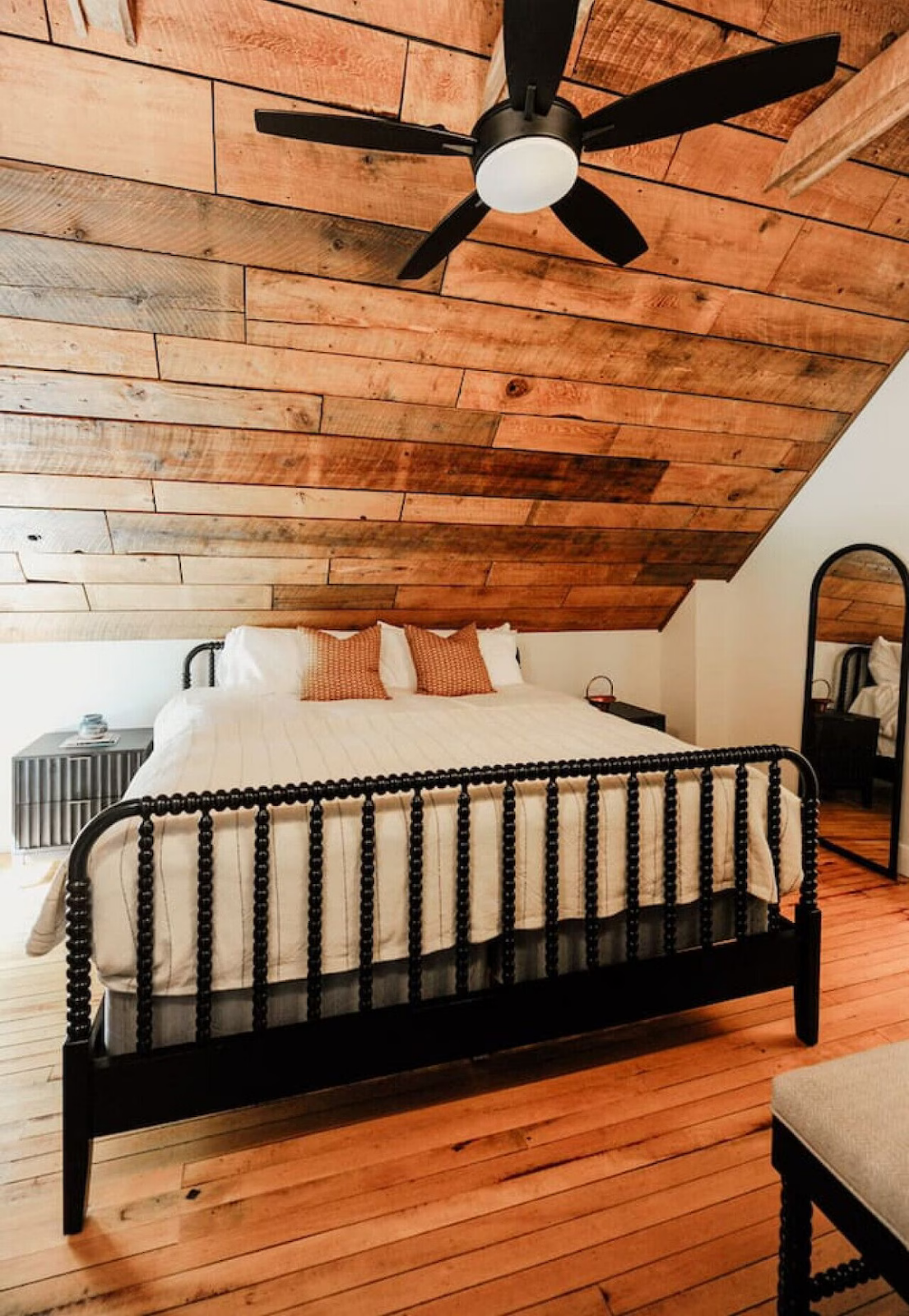 Bedroom with a wooden sloped ceiling, black metal bed frame, white bedding, and three orange throw pillows. A ceiling fan is mounted above, and there are black nightstands with lamps on either side of the bed. A standing mirror is visible on the righ
