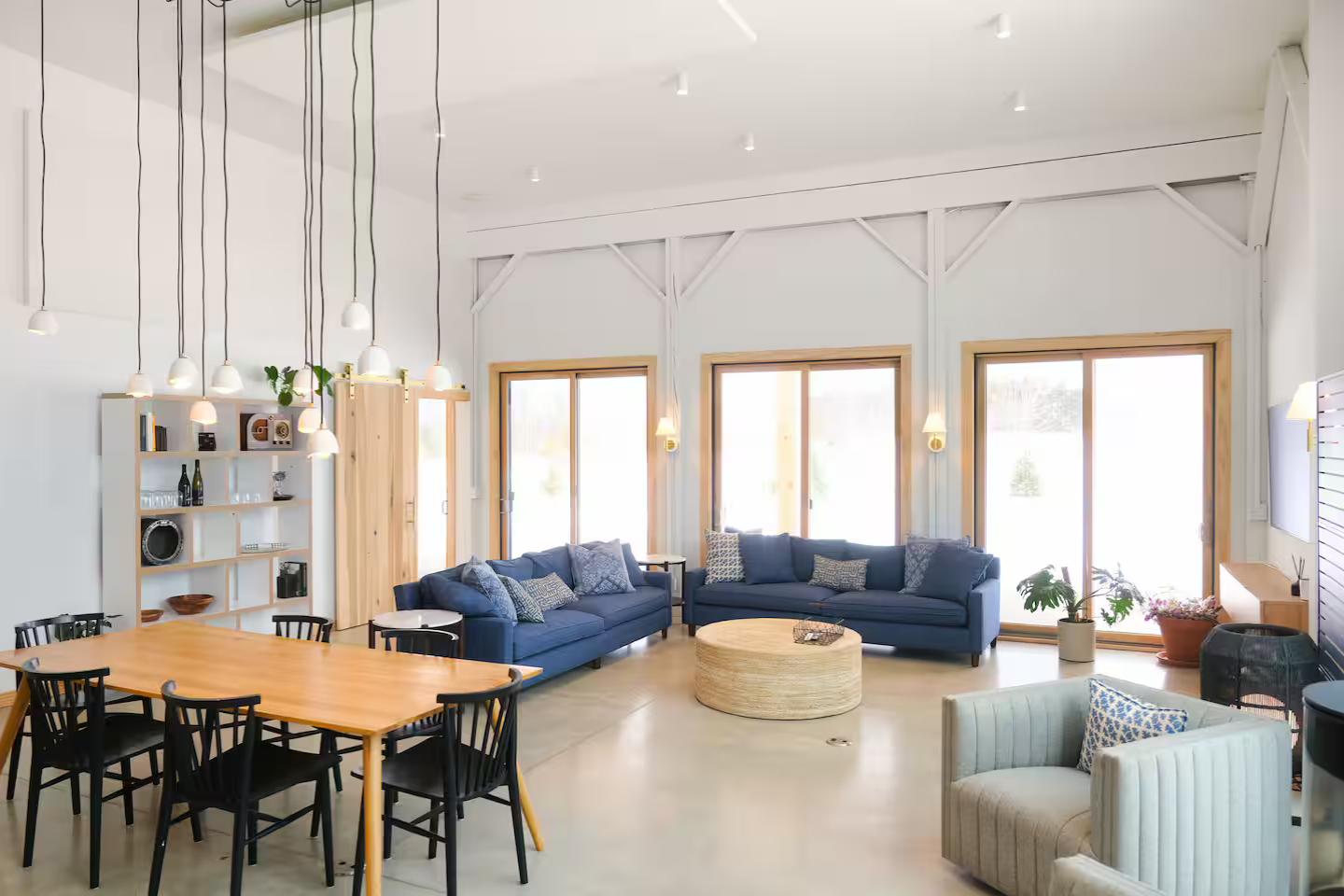 A spacious living room with white walls, large windows, and wooden accents, furnished with blue sofas, a round beige ottoman, a dining table with black chairs, and decorative plants.