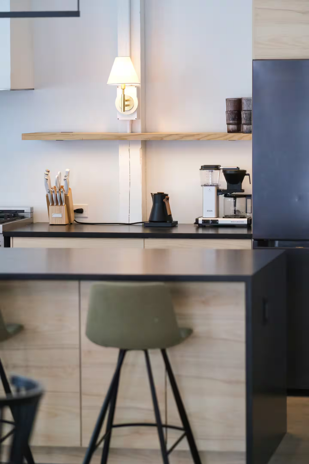 Modern kitchen with black countertop, wooden cabinets, and minimalist decor, including a lamp on a floating shelf, a knife block, a coffee machine, and an electric kettle.