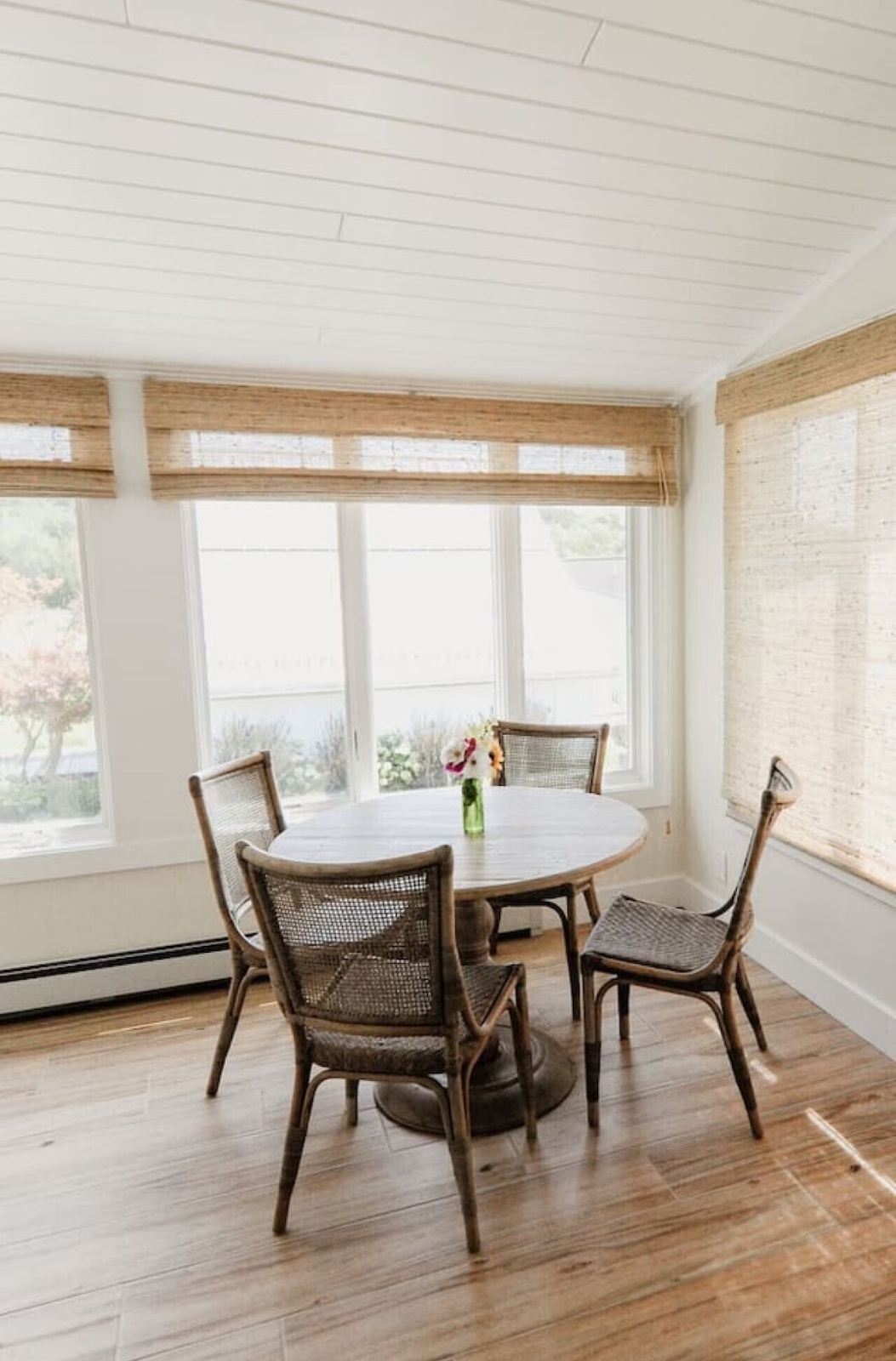 A small round dining table with four wicker chairs and a small green vase with flowers, in a bright sunlit room with large windows and wooden floors.