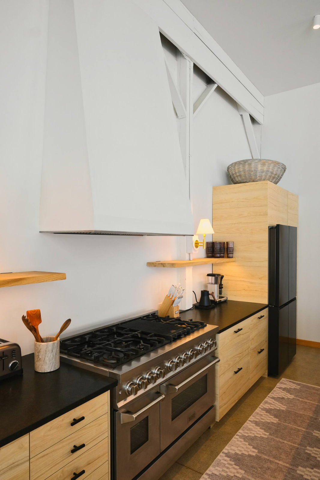Modern kitchen with black countertops, light wood cabinets, a stainless steel stove, and a refrigerator. There are small kitchen appliances, utensils, and a decorative basket on top of the cabinets. A wall-mounted light and a patterned rug are also v