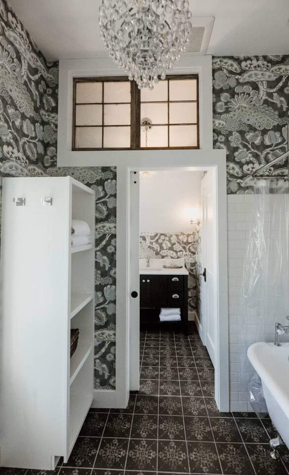 A vintage bathroom with black and white floral wallpaper, a crystal chandelier, a white shelving unit with towels, a white subway tile wall, black vanity with towel, and patterned black and white floor tiles.