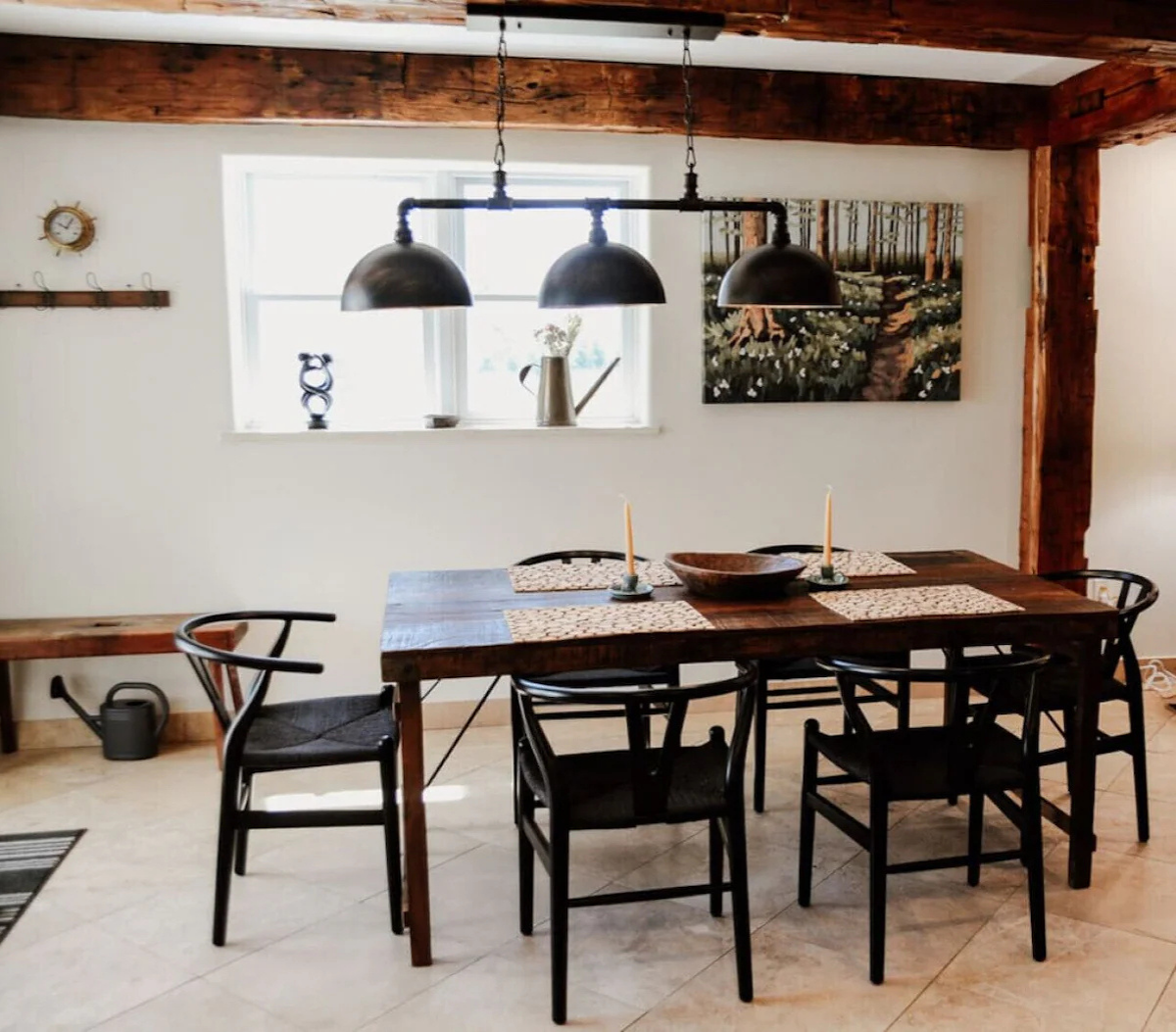 Dining room with wooden table, black chairs, black pendant light fixture, window with window sill and plant, wall art of forest scene, clock on wall, watering can on floor, and bench along wall.