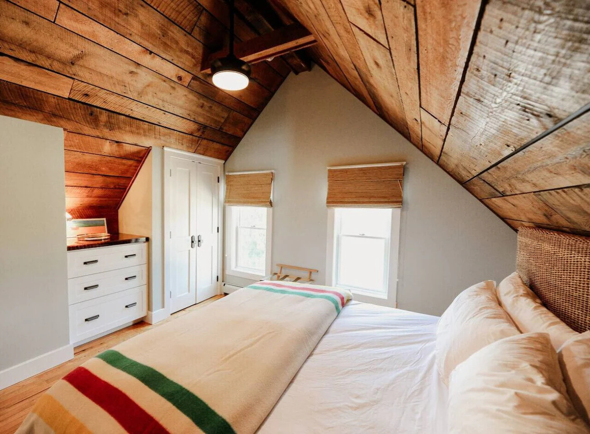 A cozy bedroom with a sloped wooden ceiling, featuring a bed with beige pillows and a cream-colored blanket with multicolored stripes, two windows with bamboo shades, a white dresser, and a closet door.