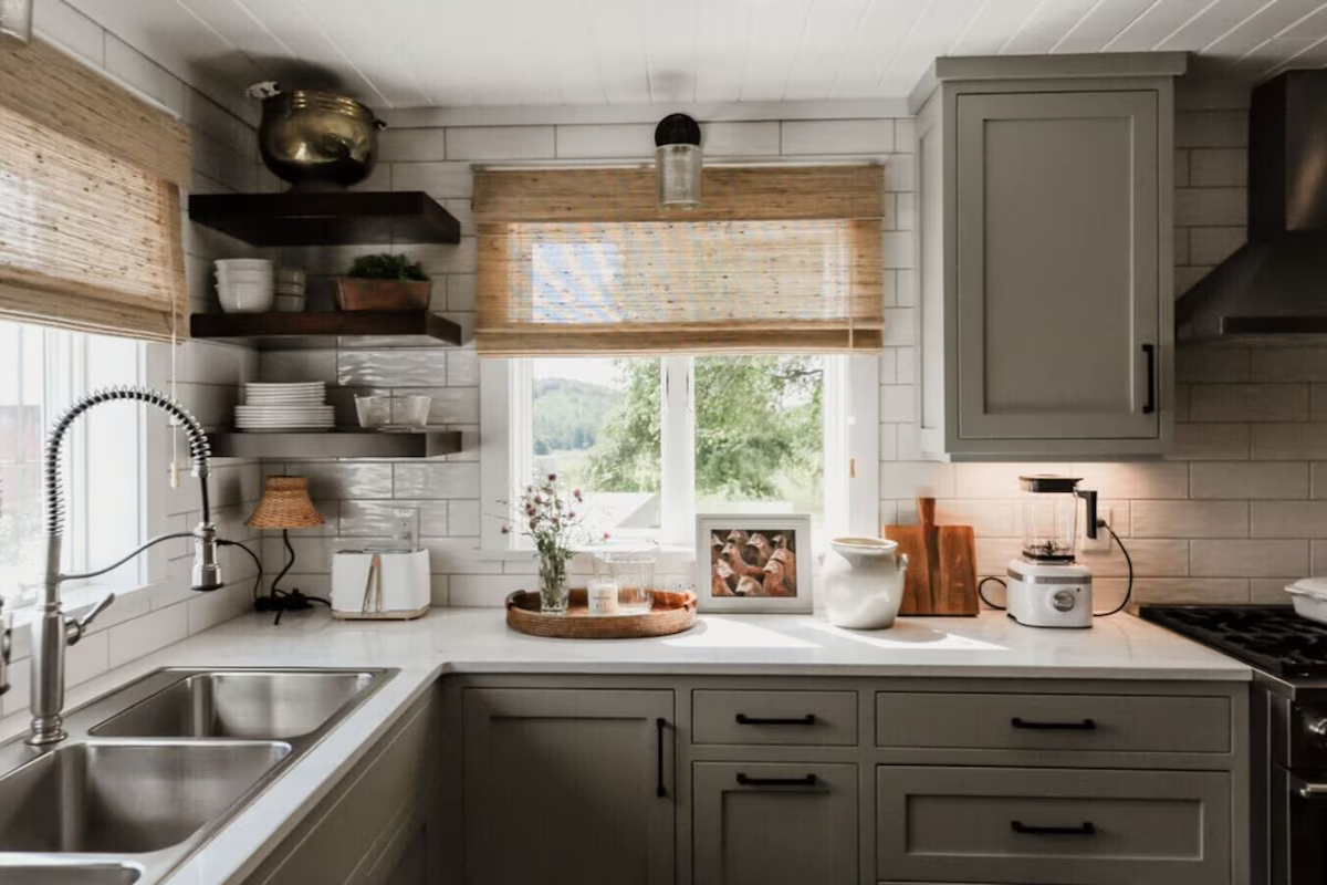 A bright kitchen with white subway tile backsplash, grey cabinets, open shelves with dishes, a running faucet over a double sink, a window with bamboo shades, and various kitchen decor items including a tray with candles, a framed photo, and a toaste