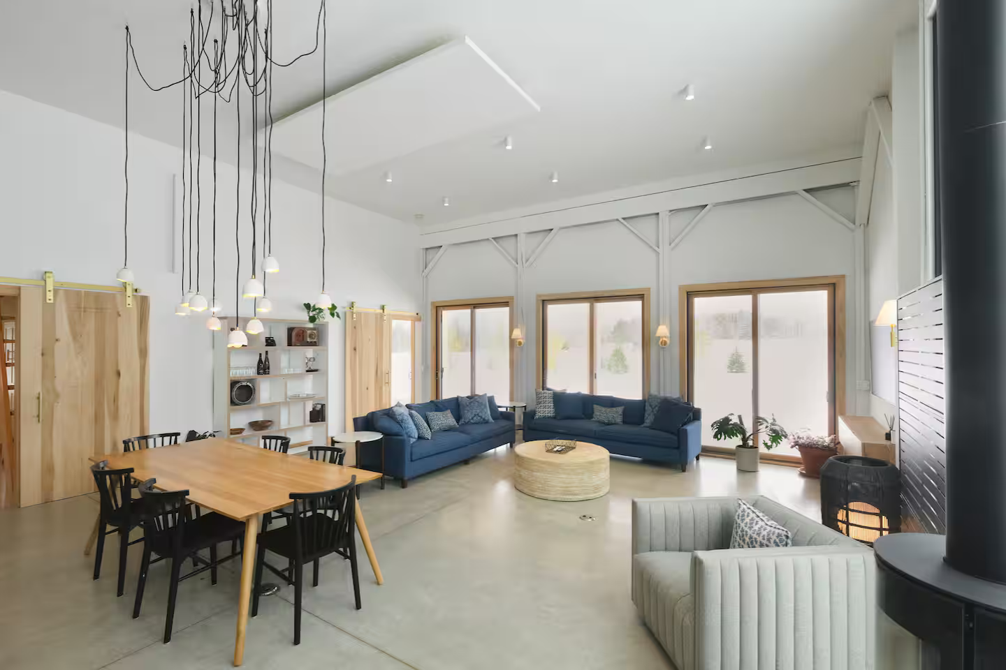Spacious living room with high vaulted ceiling, three large windows, two navy sofas with pillows, white armchair, round beige coffee table, wooden dining table with black chairs, white bookshelf, hanging lights, potted plants, and minimal decor.