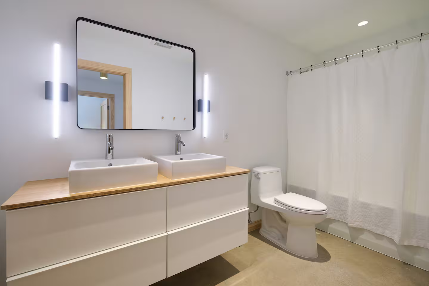 Modern bathroom with white walls, double vessel sinks on a wooden vanity, large mirror, wall-mounted lighting, a toilet, and cream-colored curtains.